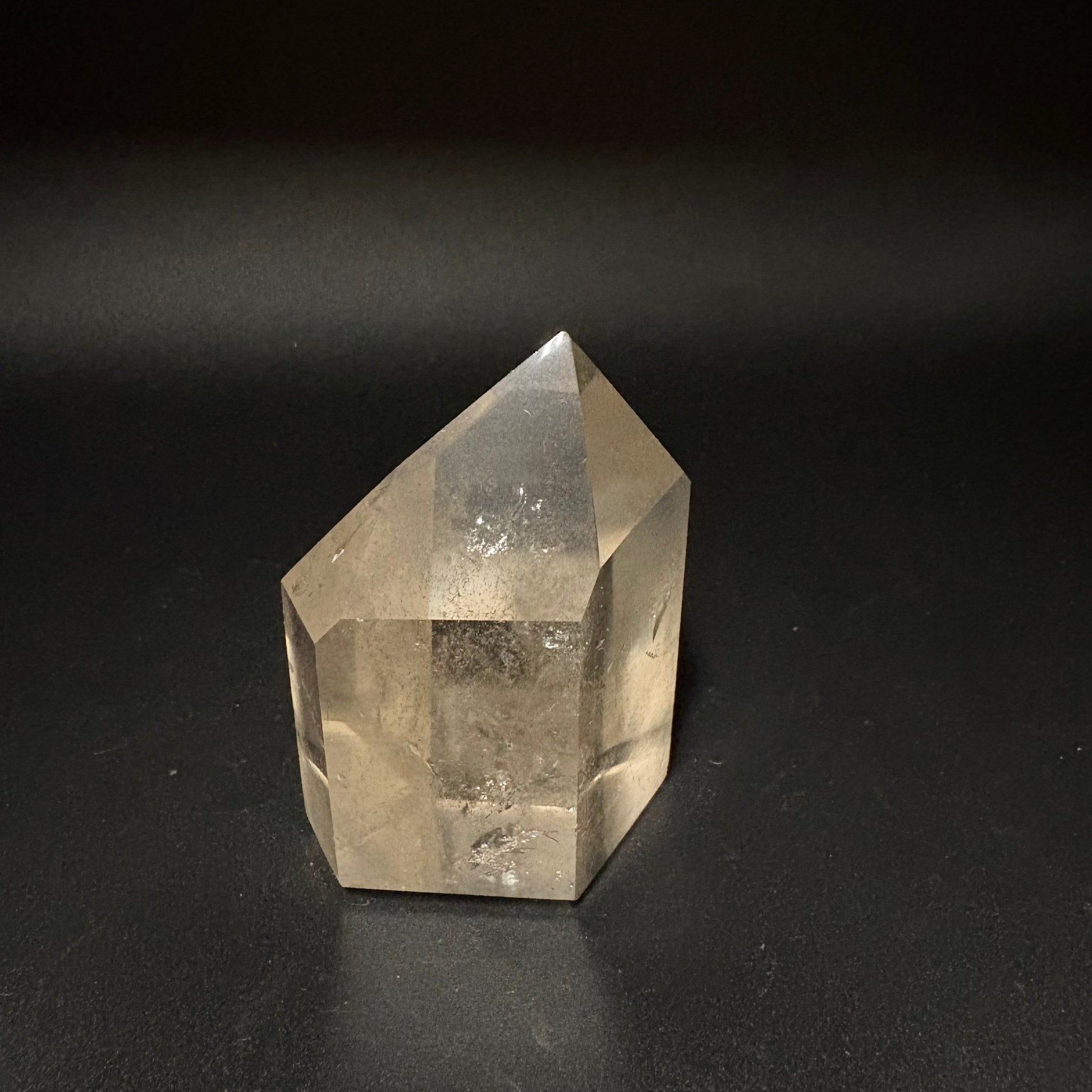 A natural clear quartz crystal point with internal inclusions, standing on a dark black surface against a dark background.