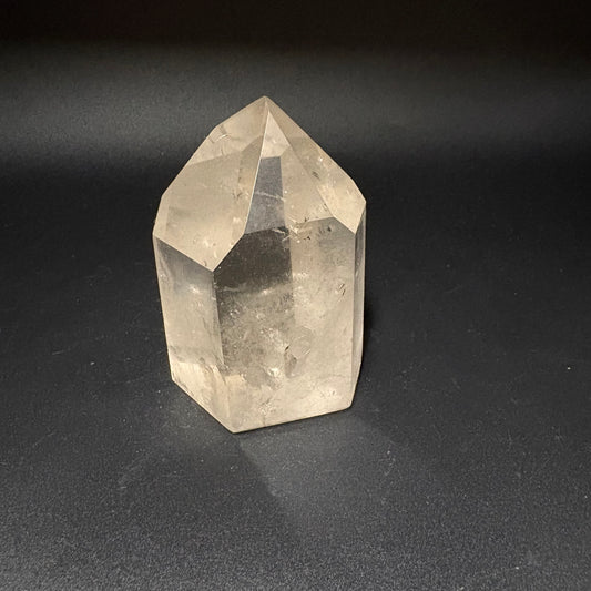 A natural clear quartz crystal point with geometric facets, standing upright on a dark gray textured surface.