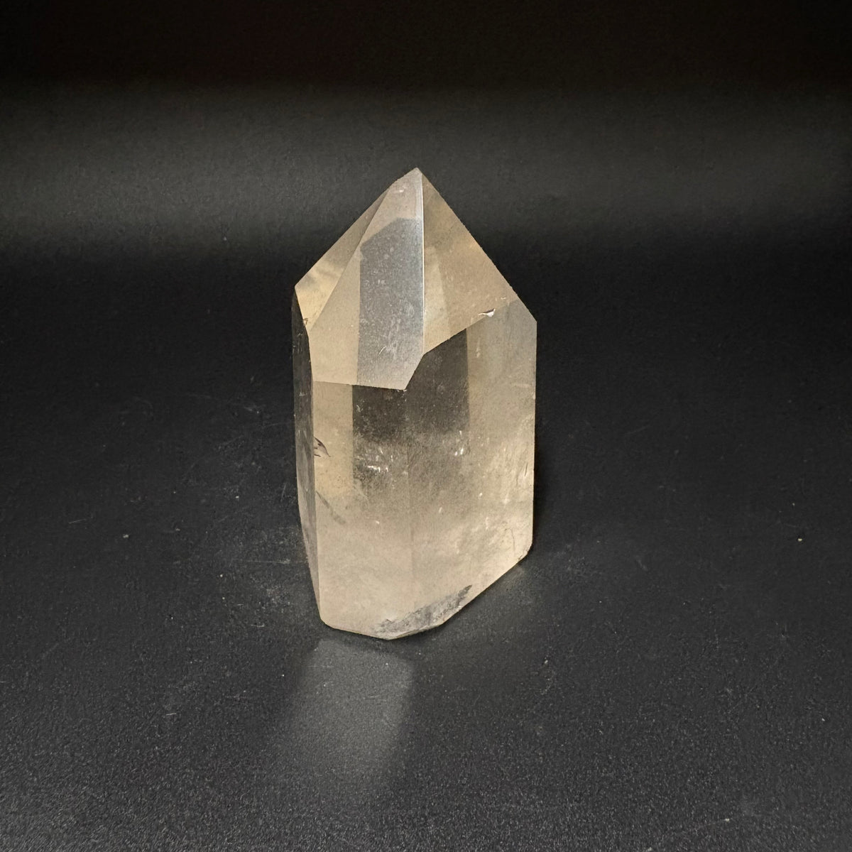 A natural clear quartz crystal point with a pointed top and hexagonal base, standing upright on a dark, textured surface.