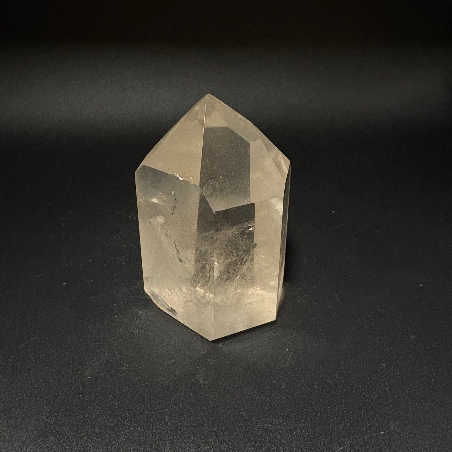A natural clear quartz crystal point with geometric facets, standing upright on a dark, textured surface.