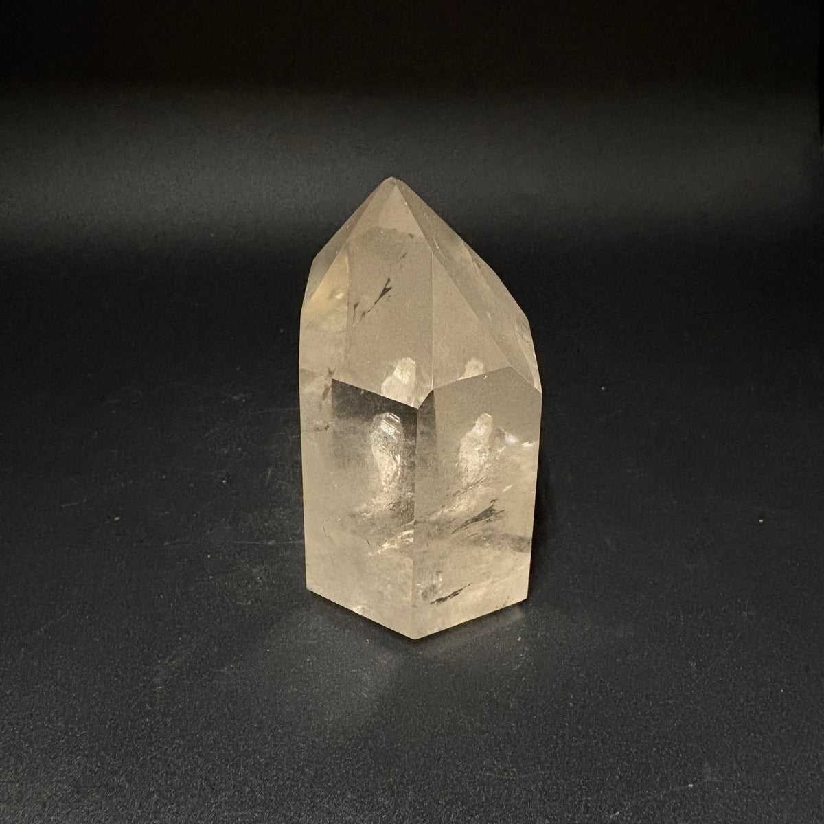 A natural clear quartz crystal point with internal inclusions, standing upright on a dark black background.