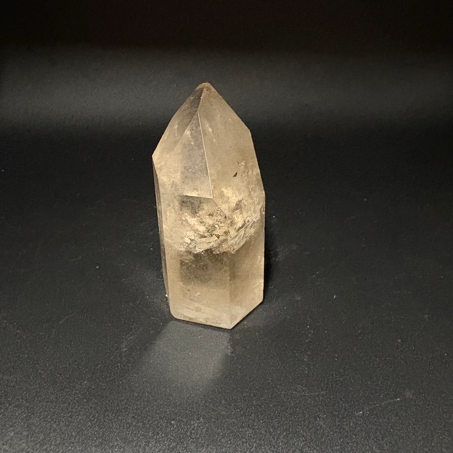 A natural clear quartz crystal point with internal inclusions, standing upright on a dark gray surface.