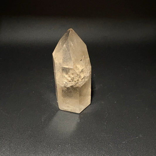 A natural clear quartz crystal point with internal inclusions, standing upright on a dark gray surface.