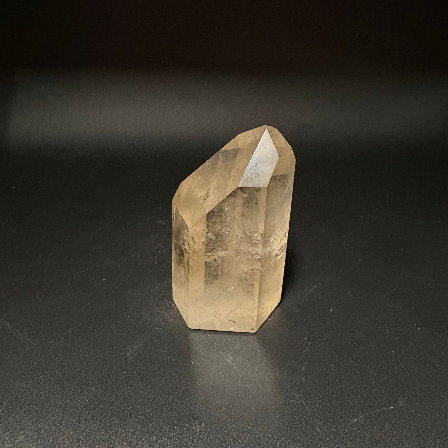 A natural citrine crystal point with a hexagonal shape, showing a warm yellowish-brown translucent color, standing on a dark gray surface against a dark background.