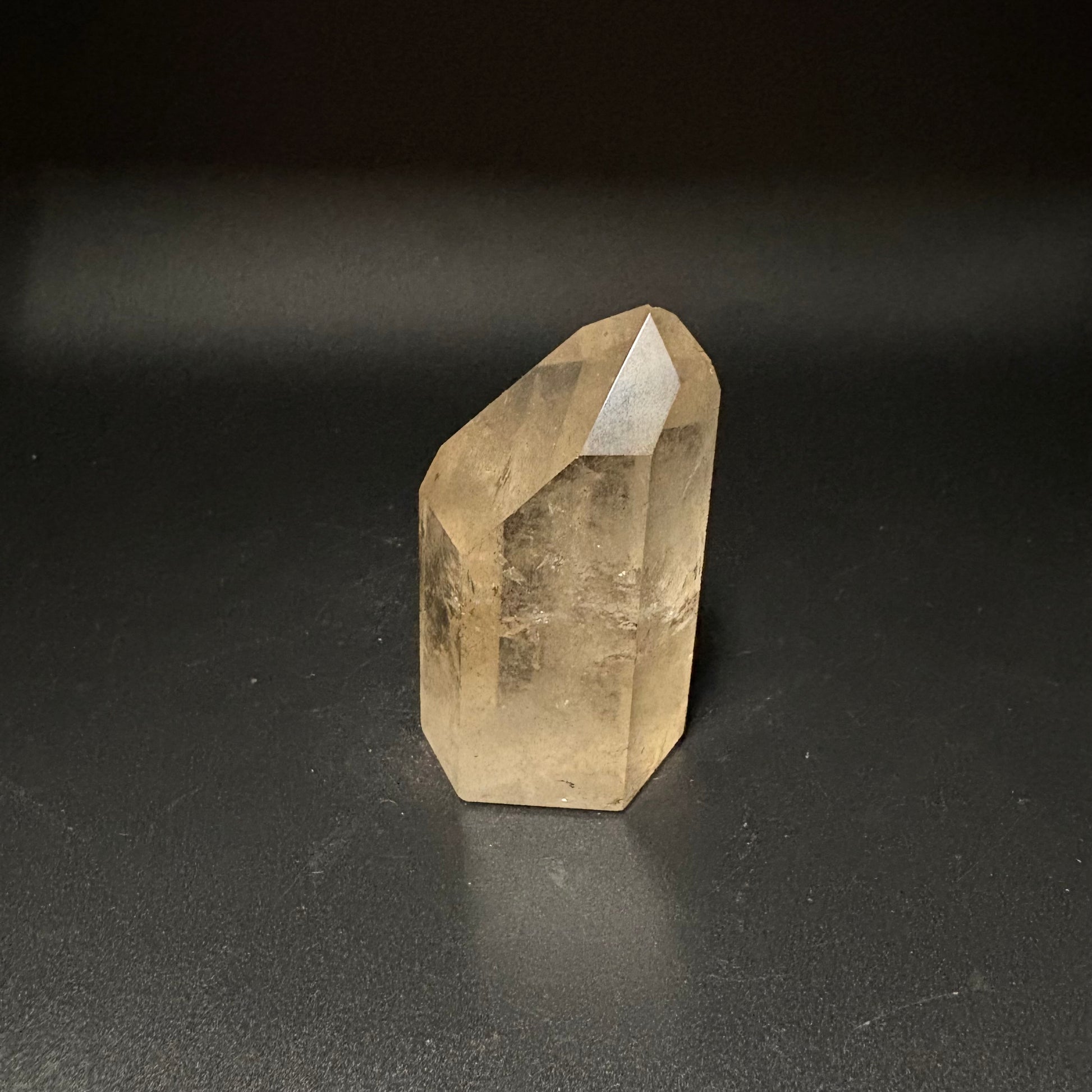 A natural citrine crystal point with a hexagonal shape, showing a warm yellowish-brown translucent color, standing on a dark gray surface against a dark background.