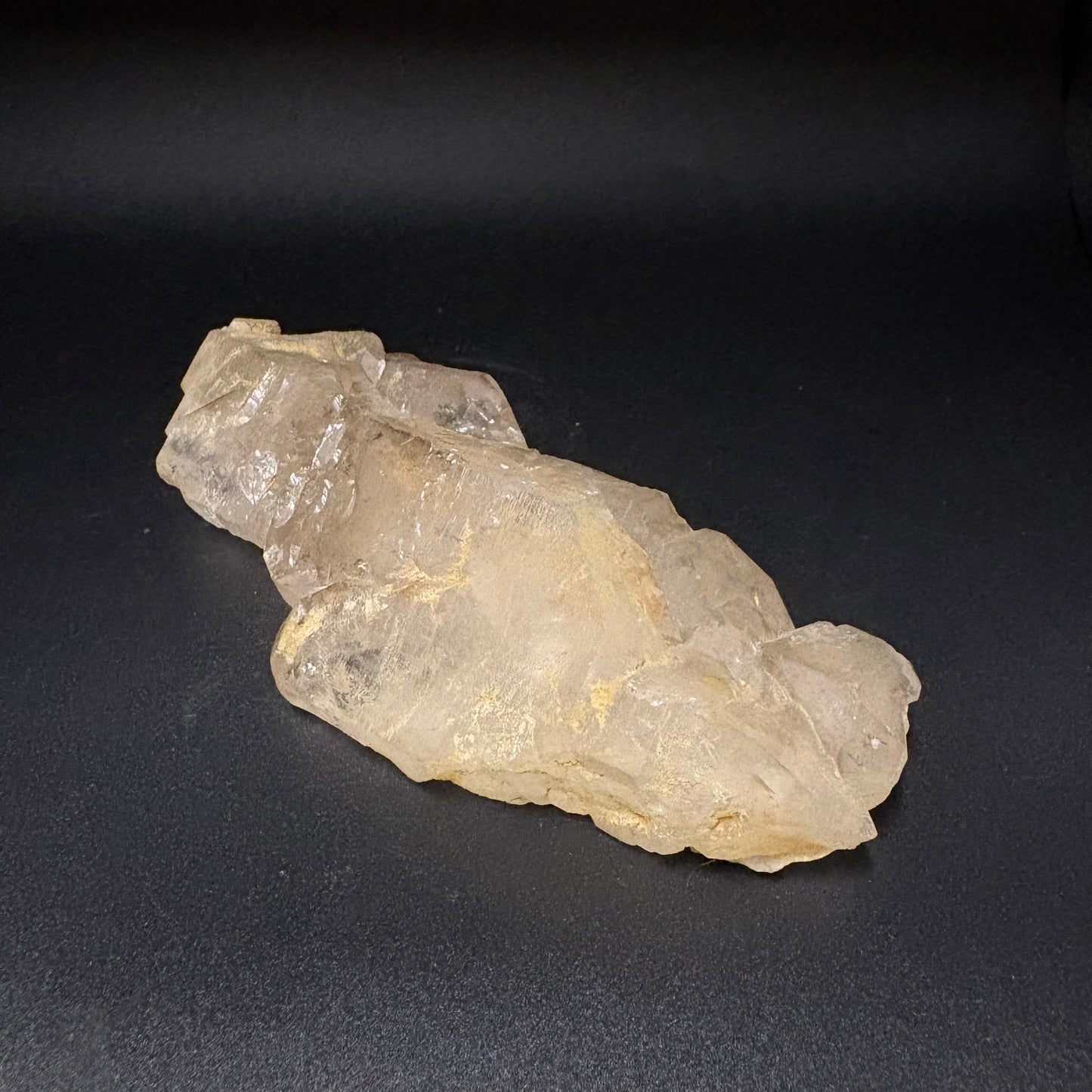 A raw, rough crystal specimen with a translucent pale yellow and beige appearance, resting on a dark textured background.