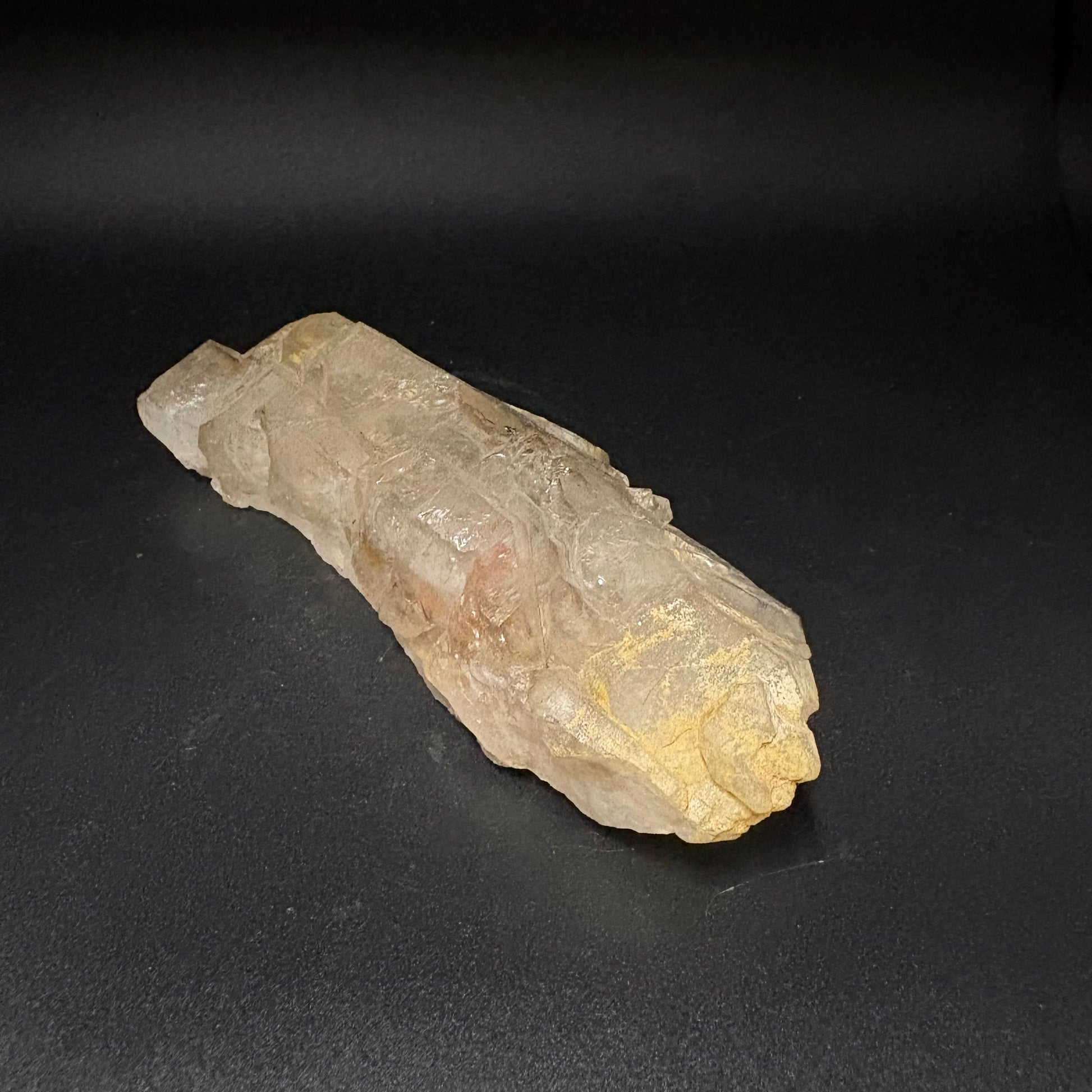 A raw, rough mineral specimen with a translucent pale yellow and beige body, displayed diagonally on a dark black background.