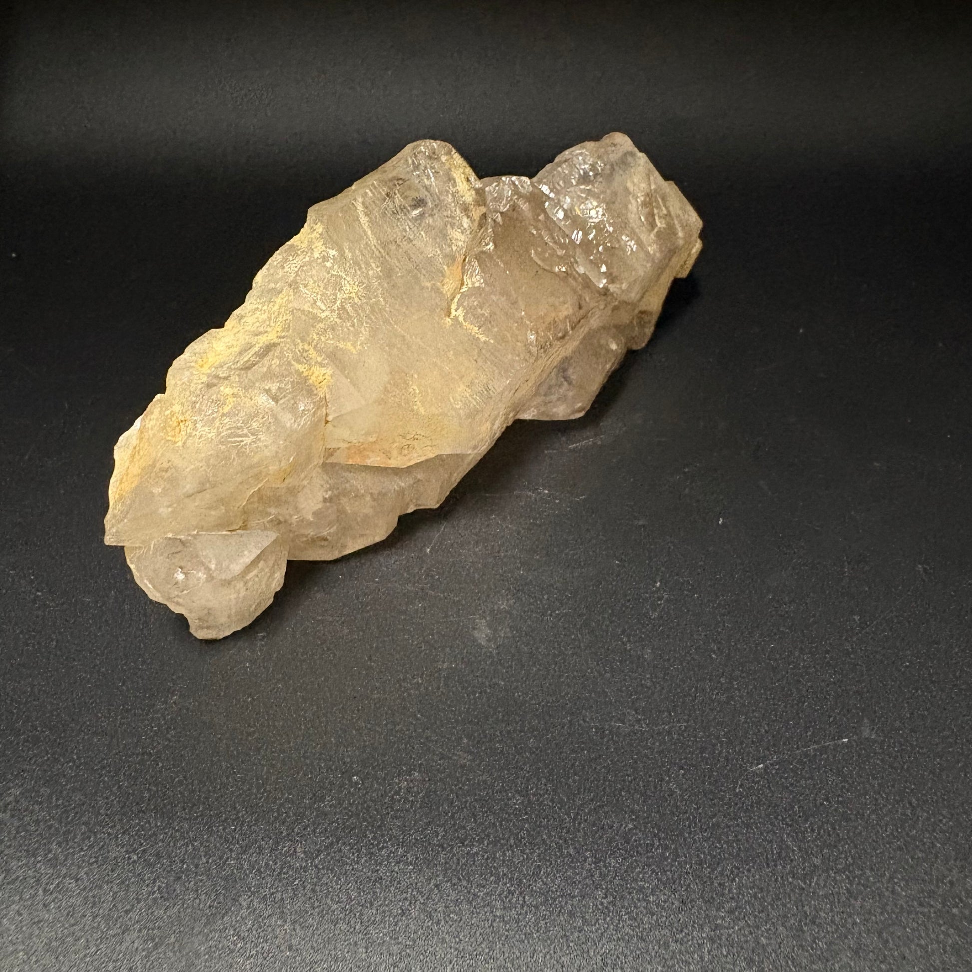 A raw, rough crystal specimen with a translucent pale yellow color and natural jagged edges, displayed on a dark gray textured surface.