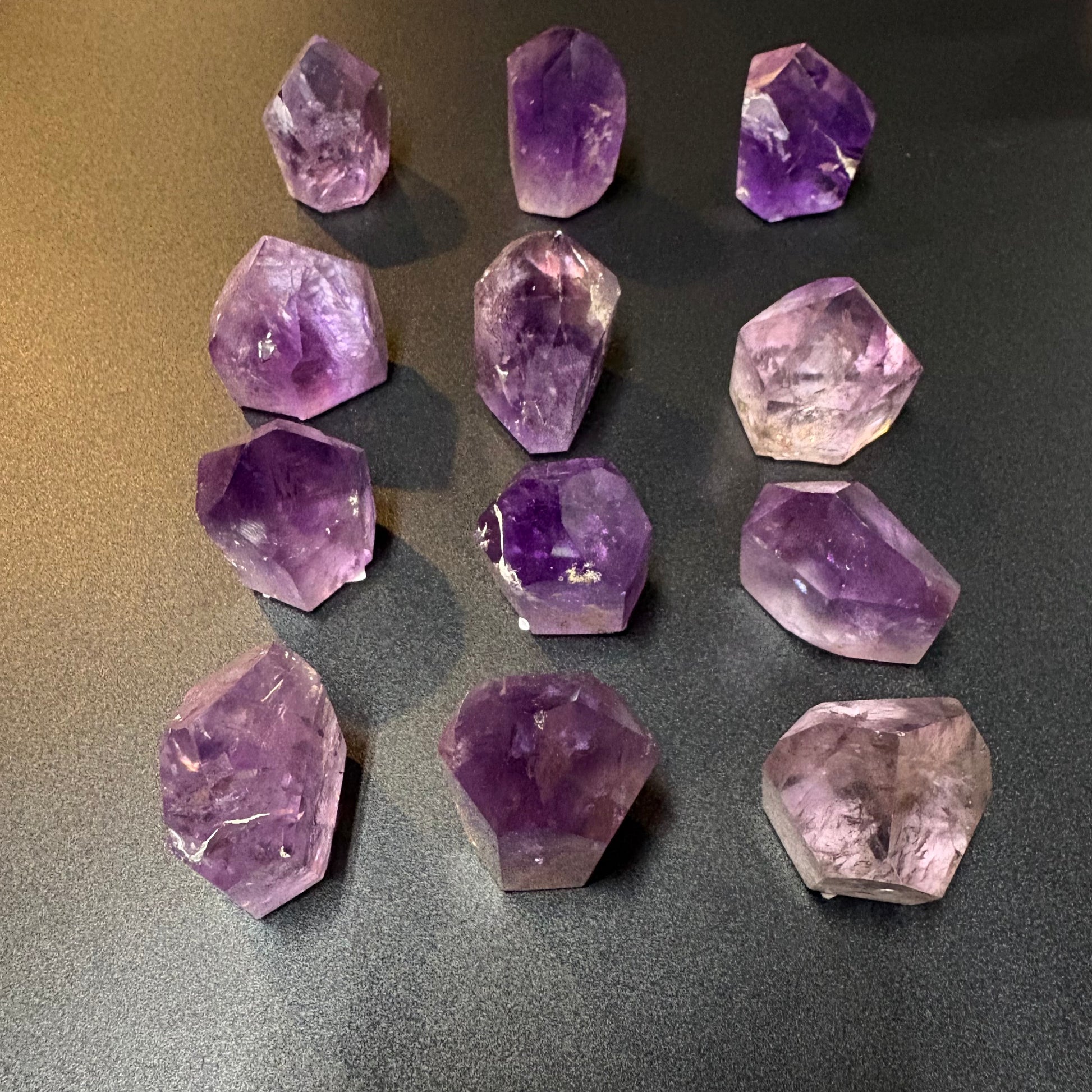 Twelve raw amethyst crystal points of various sizes arranged in a grid on a dark gray textured surface.