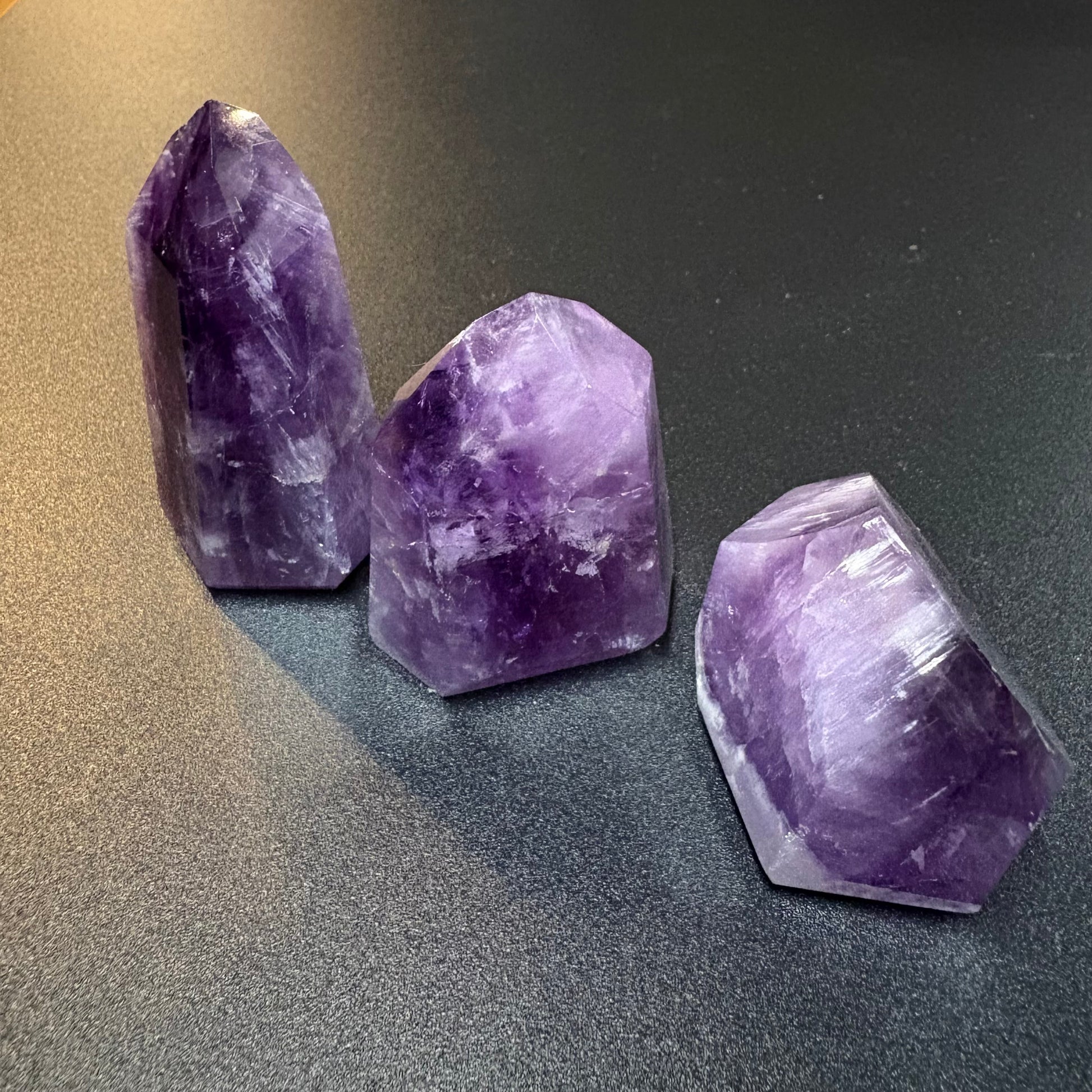 Three raw purple amethyst crystal points of different shapes standing on a dark gray textured surface.