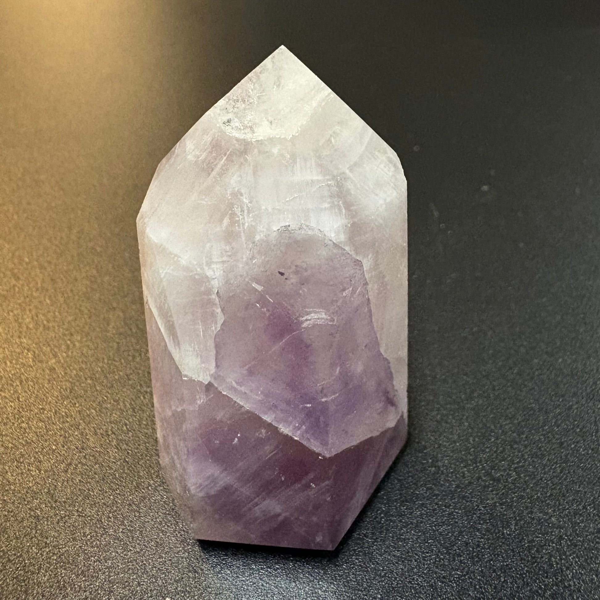 A natural amethyst crystal point tower, showcasing a gradient from translucent white at the top to deep purple at the base, resting on a dark textured surface.