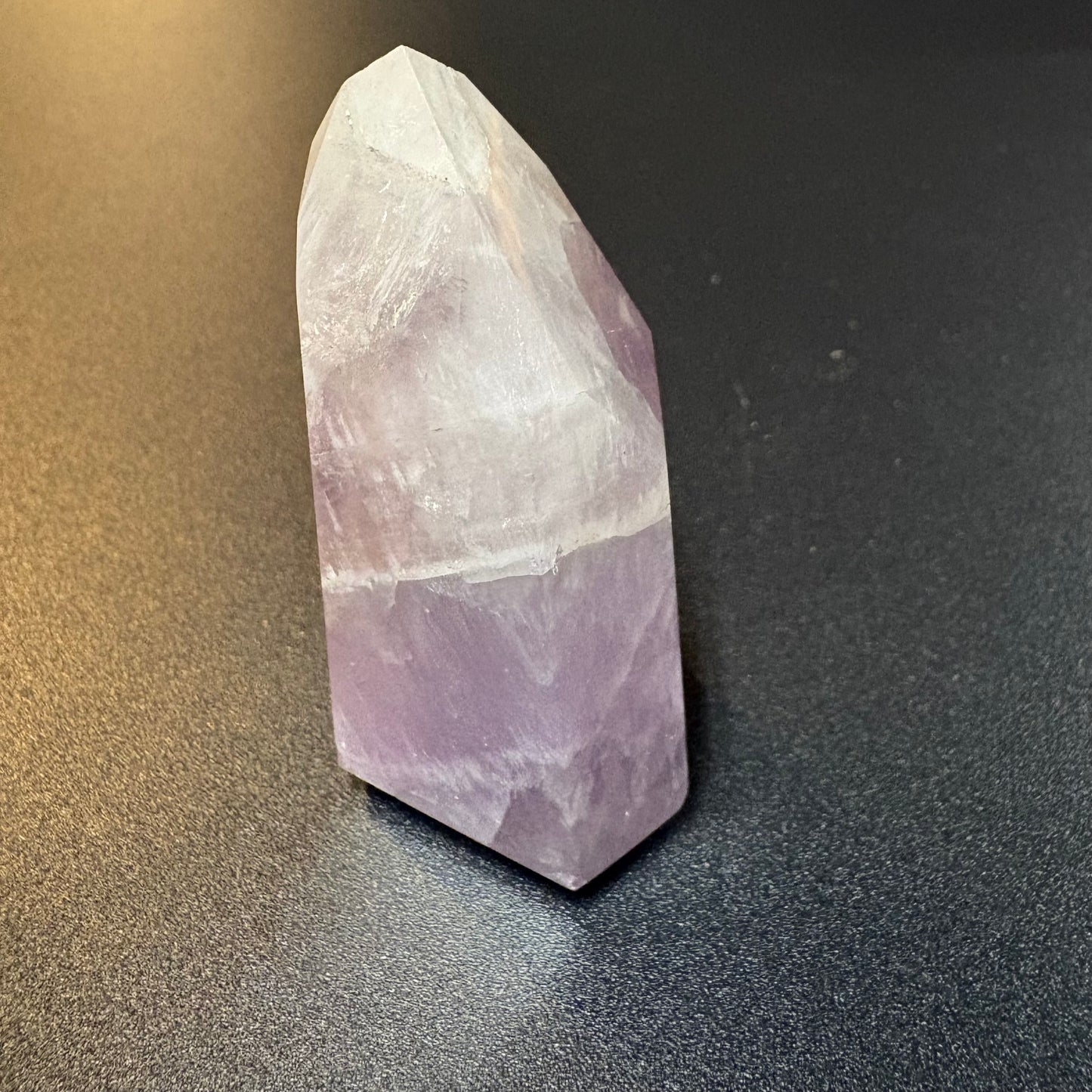 A natural amethyst crystal point tower showing a distinct white top section and a lavender-purple bottom section, standing on a dark gray textured surface.