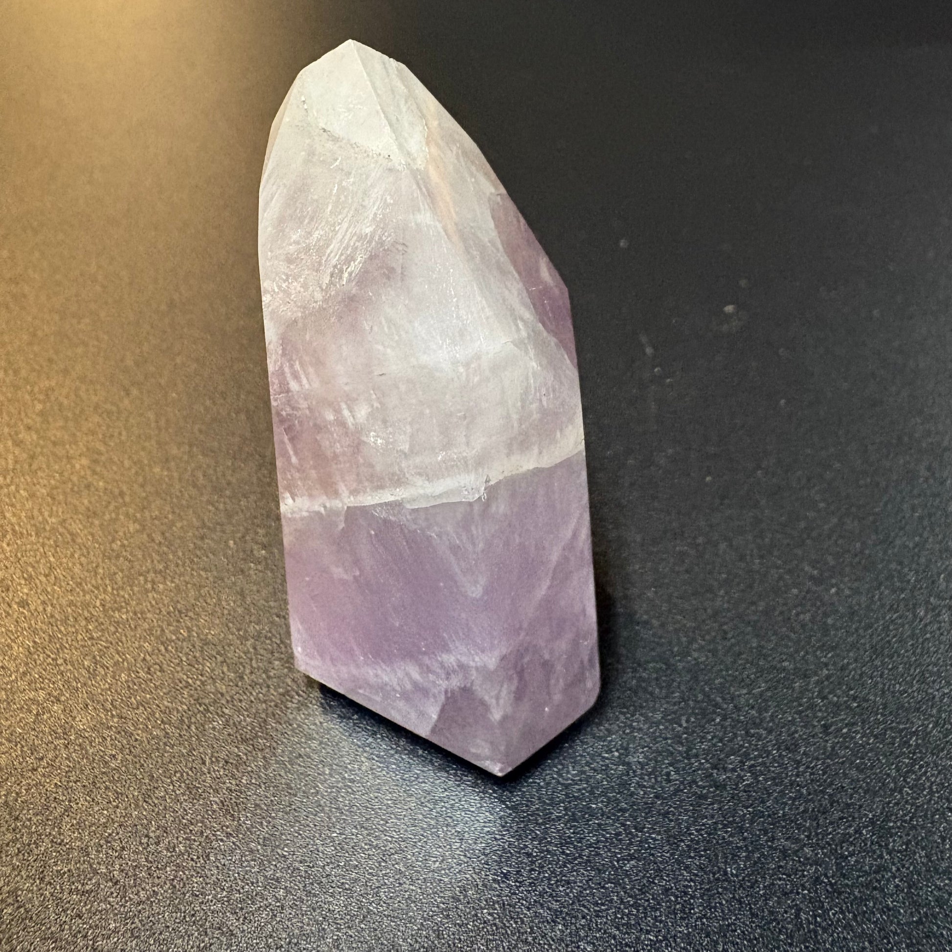 A natural amethyst crystal point tower showing a distinct white top section and a lavender-purple bottom section, standing on a dark gray textured surface.