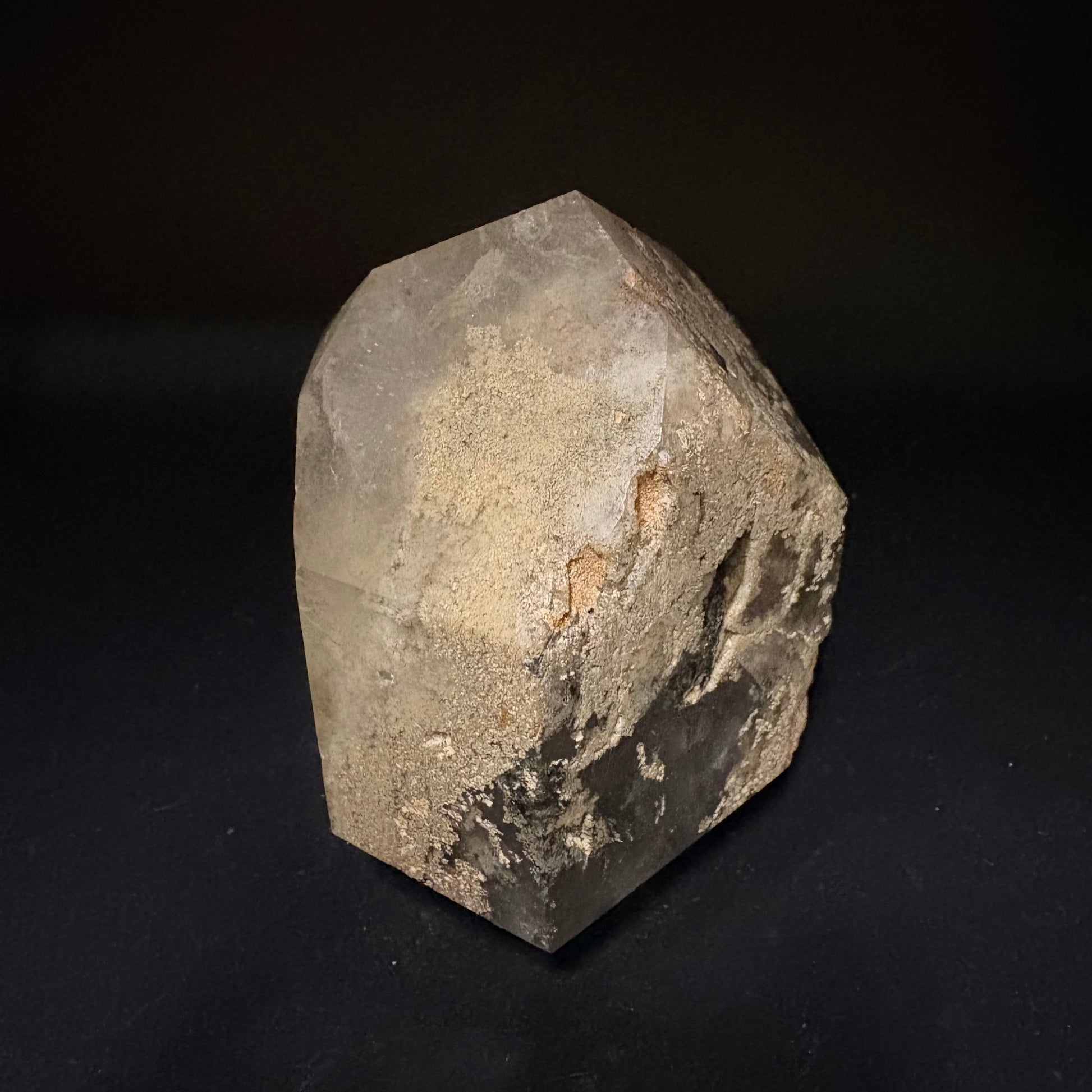 A raw, rough-cut crystal mineral specimen with natural geometric facets, showing a mix of translucent gray, sandy beige, and dark brown mineral textures, photographed on a solid black background.