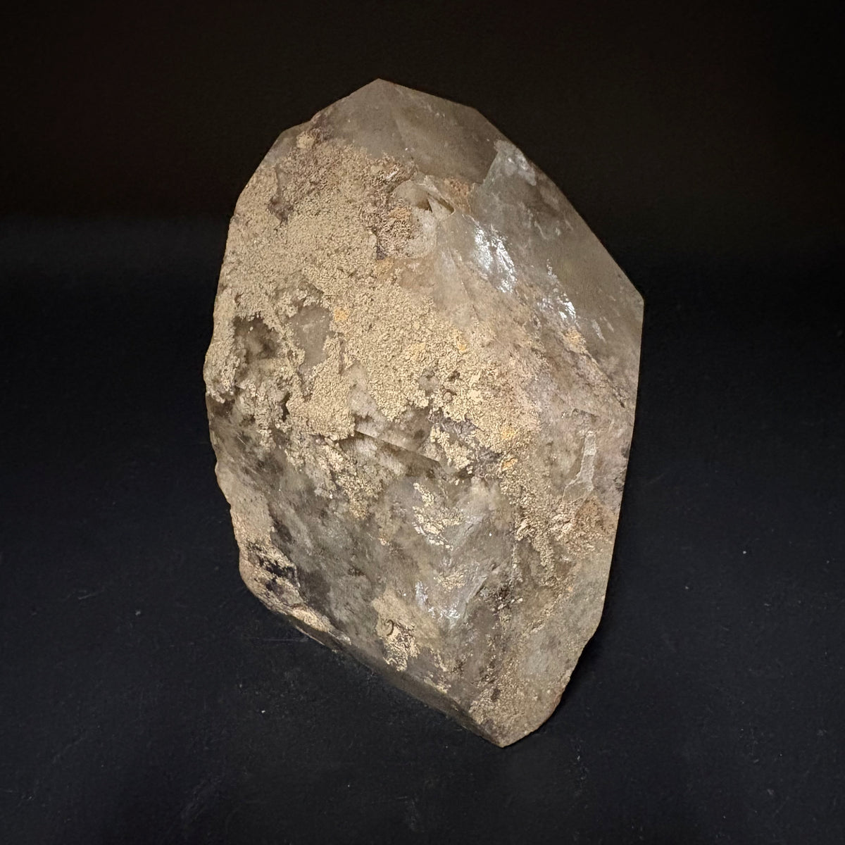 A raw, natural quartz crystal cluster with rough, earthy beige and brown mineral deposits on its surface, standing upright against a dark black background.