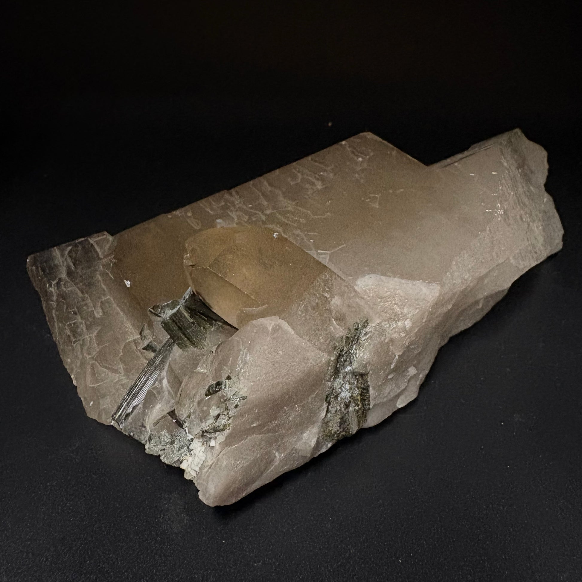 A raw mineral specimen featuring a large, translucent pale beige quartz crystal cluster with smaller metallic green and silver crystalline inclusions, displayed on a dark black background.