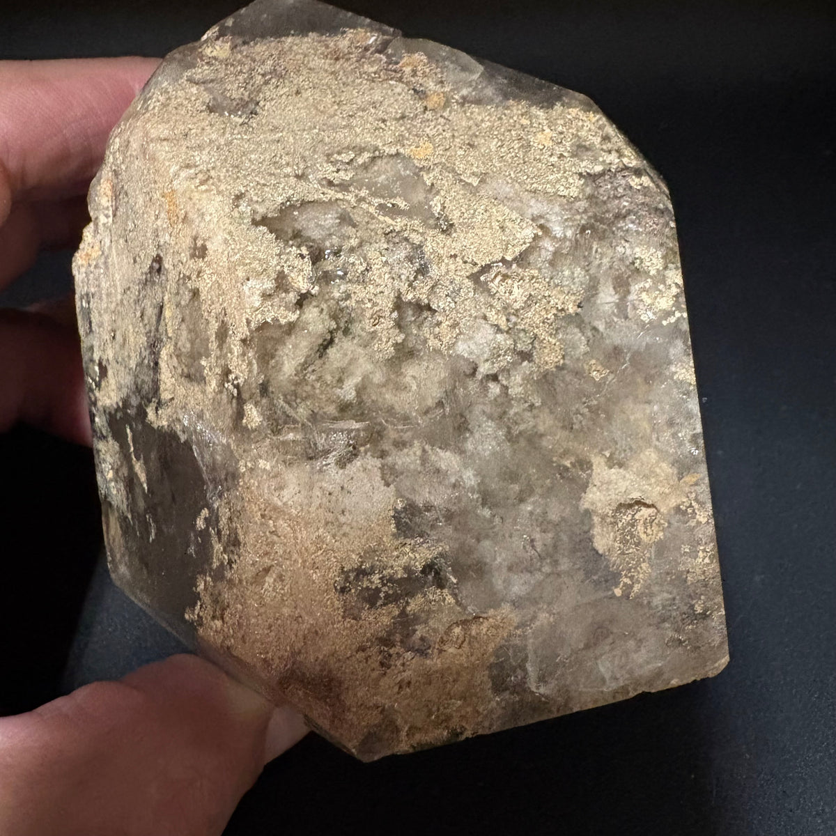 A raw, faceted crystal mineral specimen with a rough, textured surface showing natural earthy tones of beige, brown, and gray, held by a hand against a dark background.