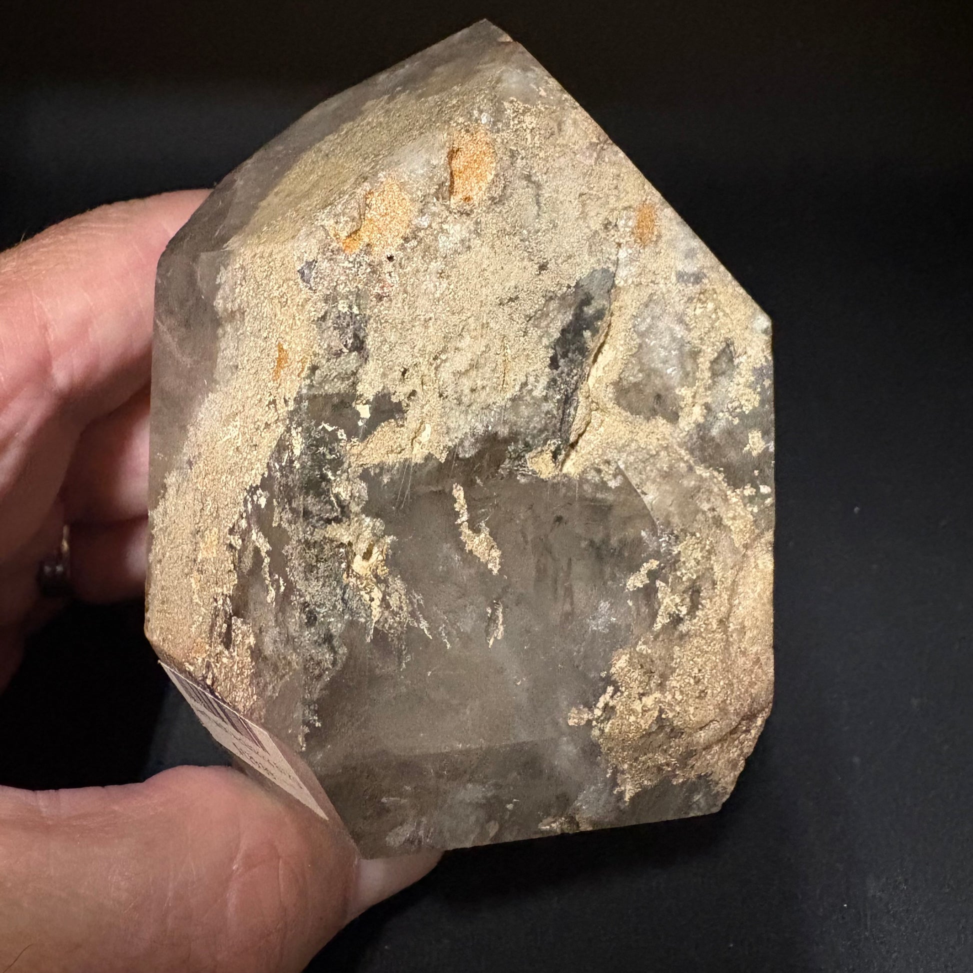 A hand holding a rough, faceted natural quartz crystal with a dusty beige and gray patina, set against a dark background.