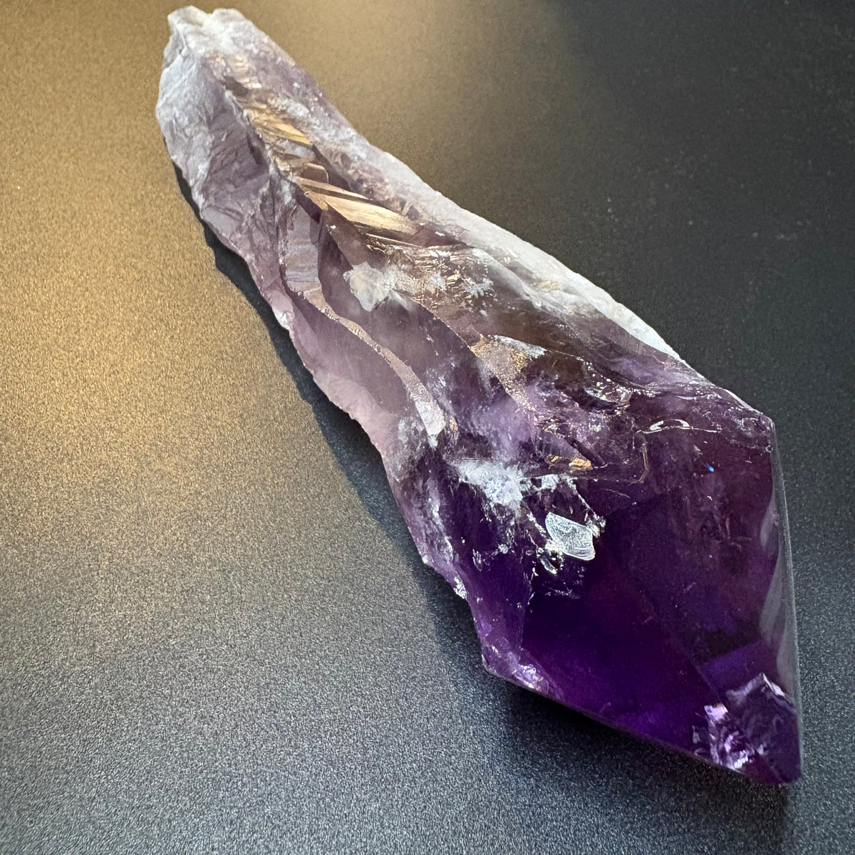 A raw amethyst crystal point with deep purple and lighter lavender tones, resting on a dark gray textured surface.