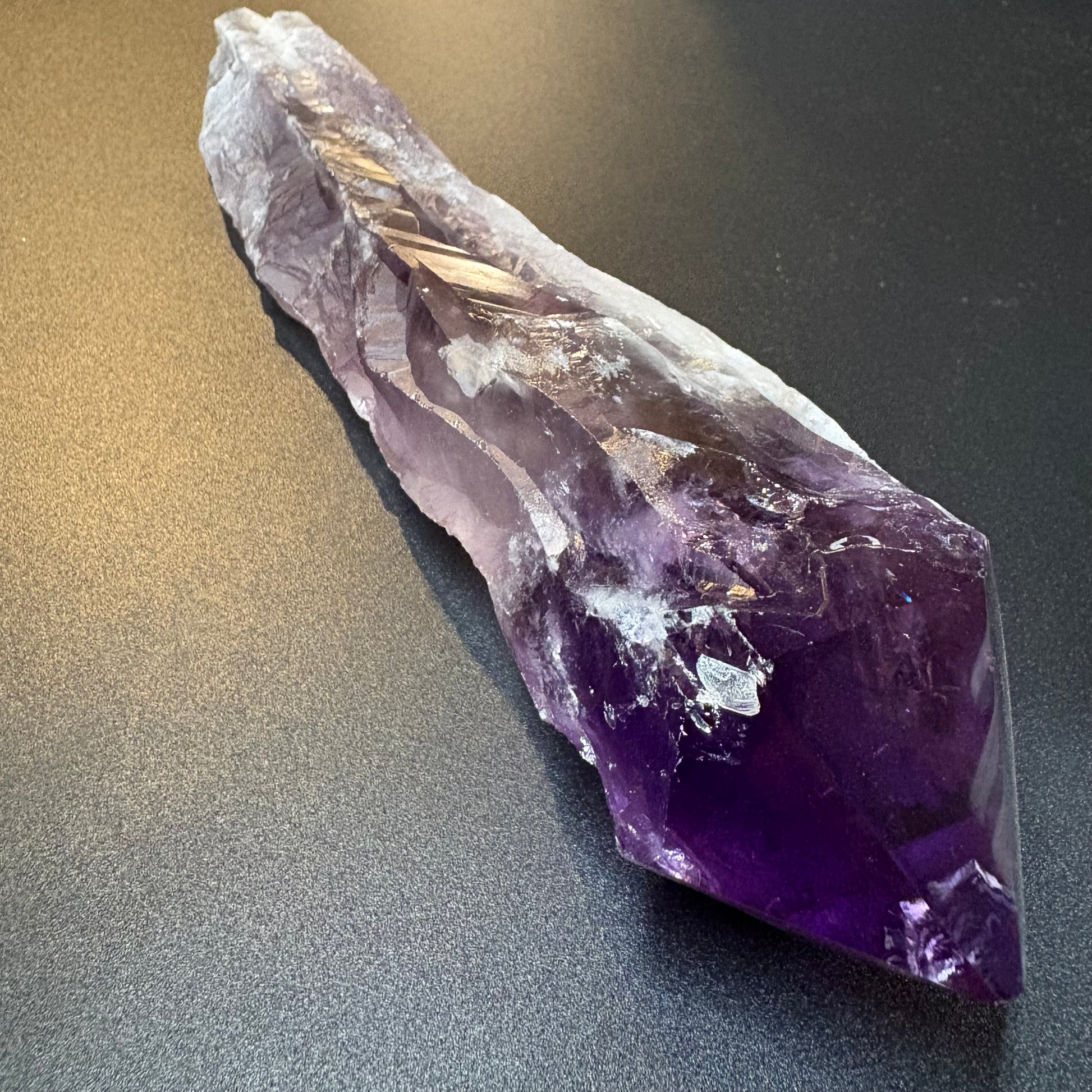 A raw amethyst crystal point with deep purple and lighter lavender tones, resting on a dark gray textured surface.