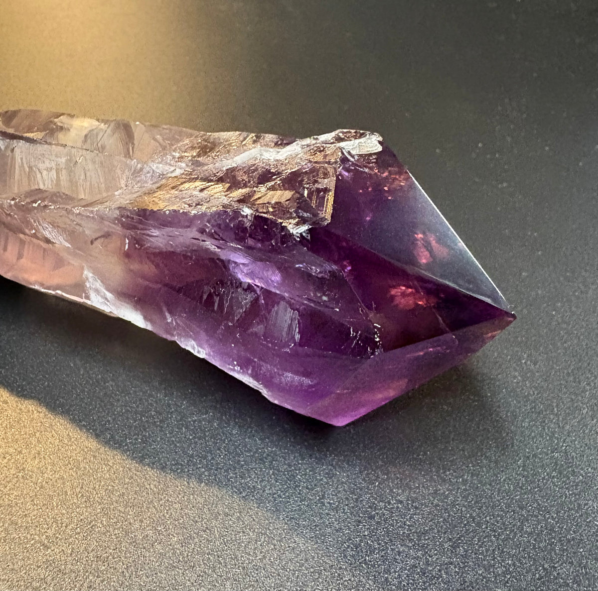 A natural amethyst crystal point with a deep purple gradient and clear outer layers, resting on a dark gray textured surface.