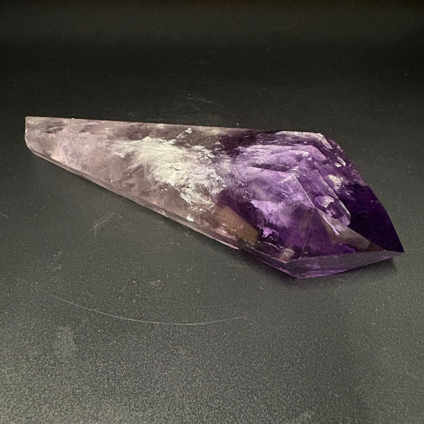 A natural amethyst crystal point with a gradient from light lavender to deep purple, resting on a dark gray surface.