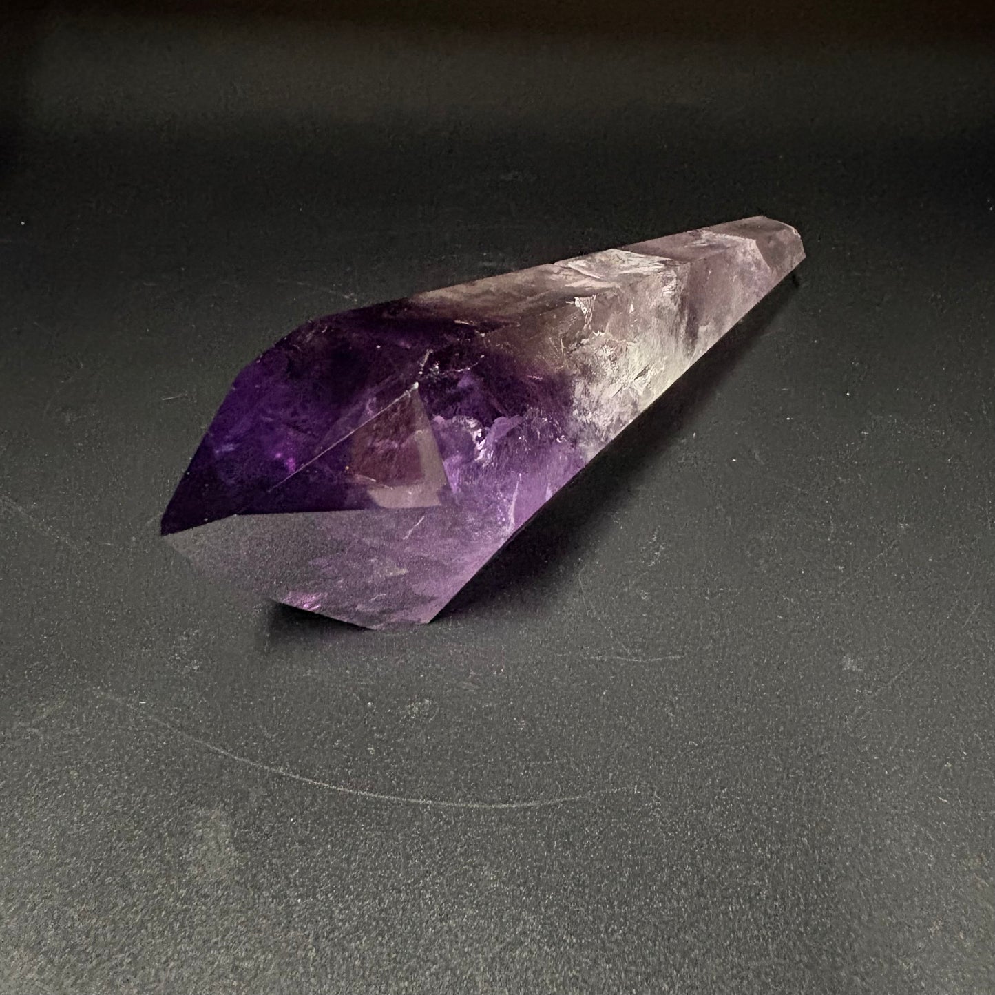 A natural amethyst crystal point with a deep purple and lavender gradient fading to translucent pale purple, resting on a dark gray surface.