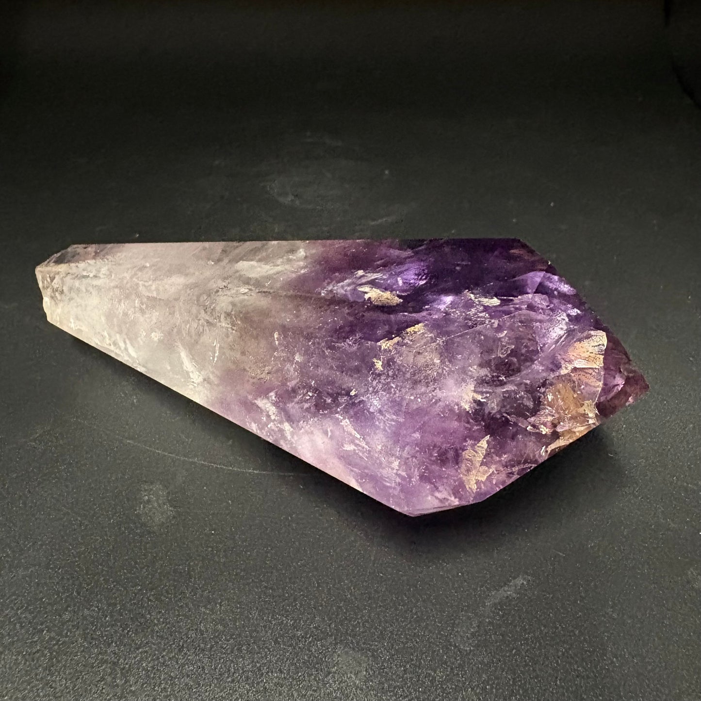 A natural amethyst crystal point with a gradient from clear and pale lavender to deep purple, resting on a dark textured surface.