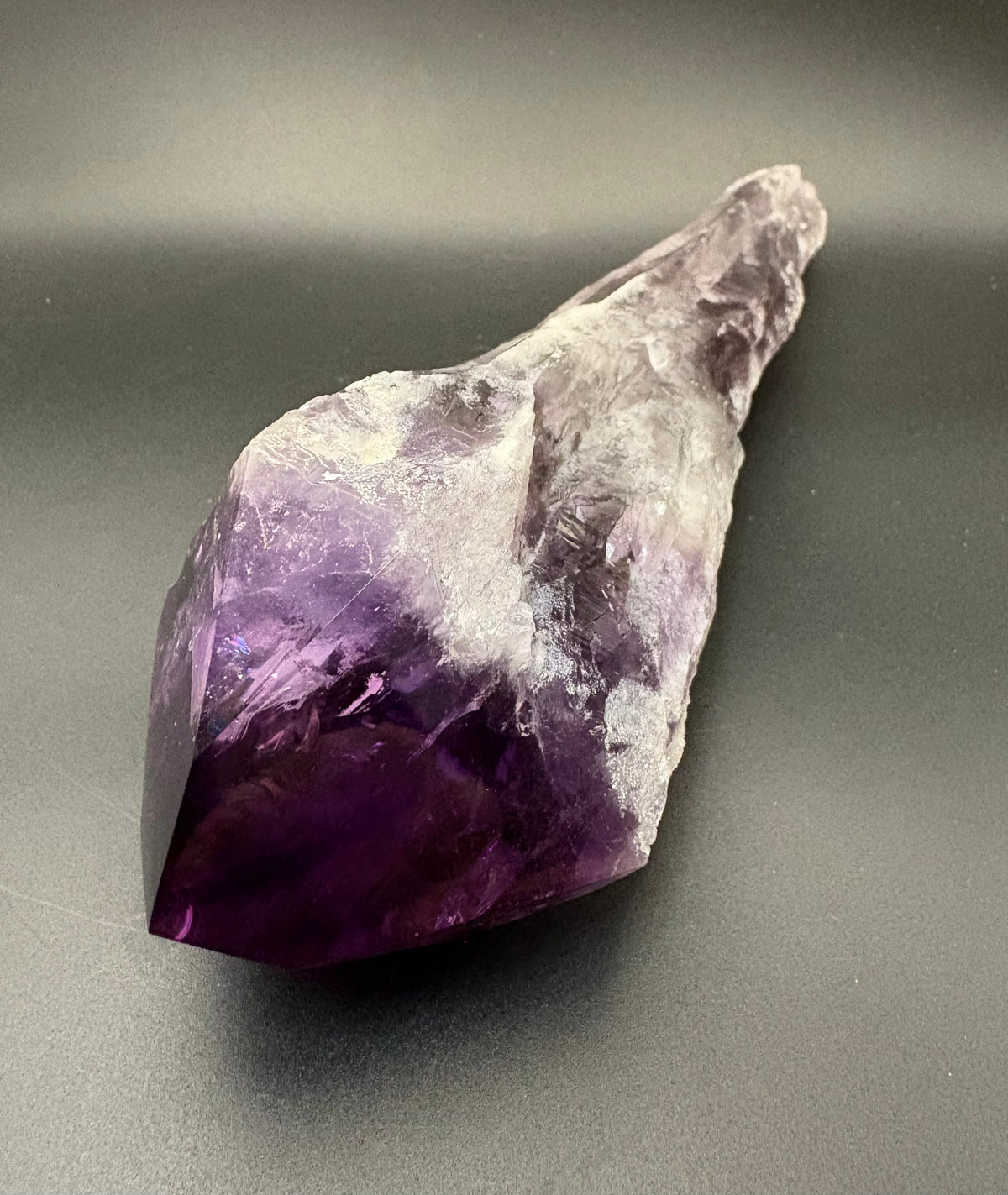 A raw amethyst crystal point with a deep purple body fading to translucent pale lavender at the tip, photographed on a dark gray background.