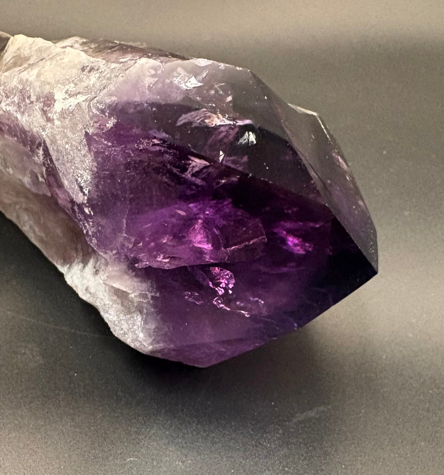 A raw amethyst crystal point showing a gradient from translucent white and light purple to deep violet, resting on a dark gray surface.