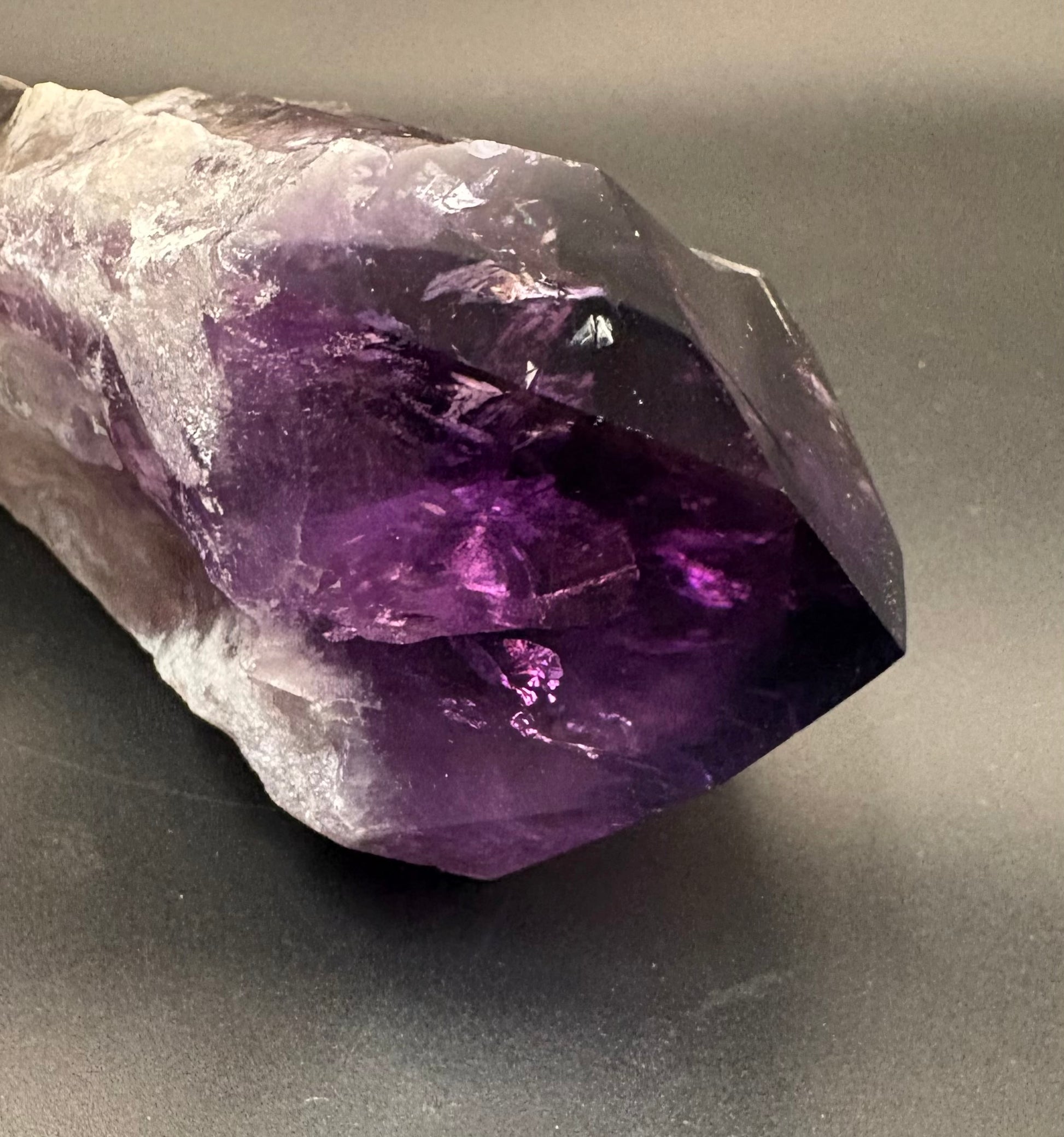 A raw amethyst crystal point showing a gradient from translucent white and light purple to deep violet, resting on a dark gray surface.