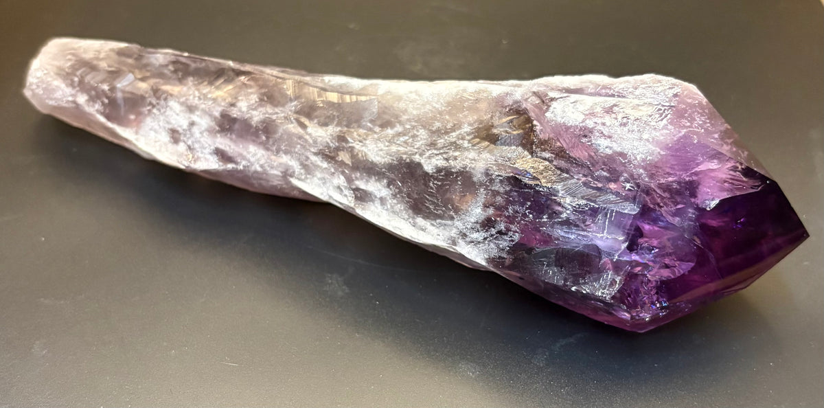 A raw amethyst crystal point with a deep purple tip and a translucent lavender body, resting on a dark gray surface.