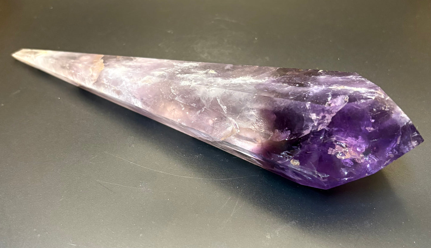 A natural amethyst crystal point with a gradient from light lavender to deep purple, resting on a dark gray surface.