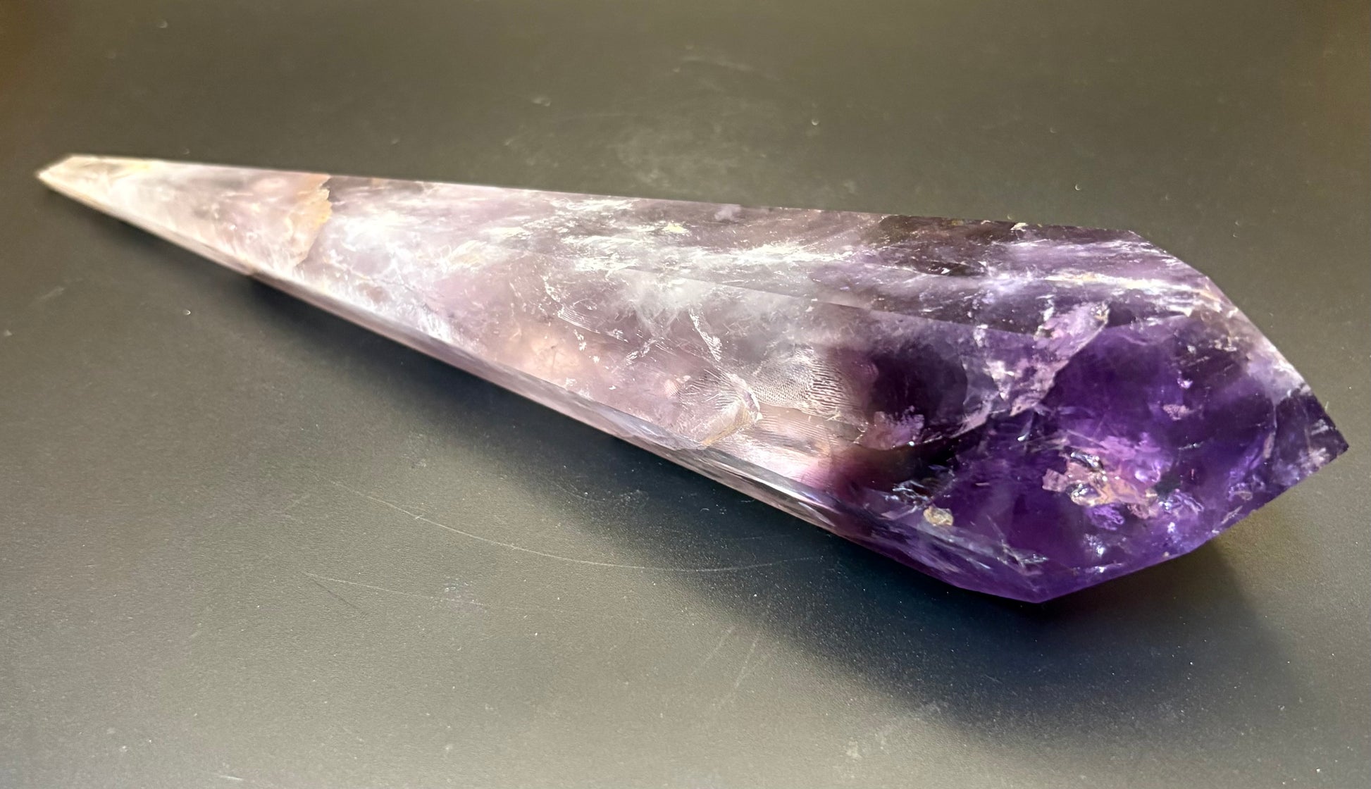 A natural amethyst crystal point with a gradient from light lavender to deep purple, resting on a dark gray surface.