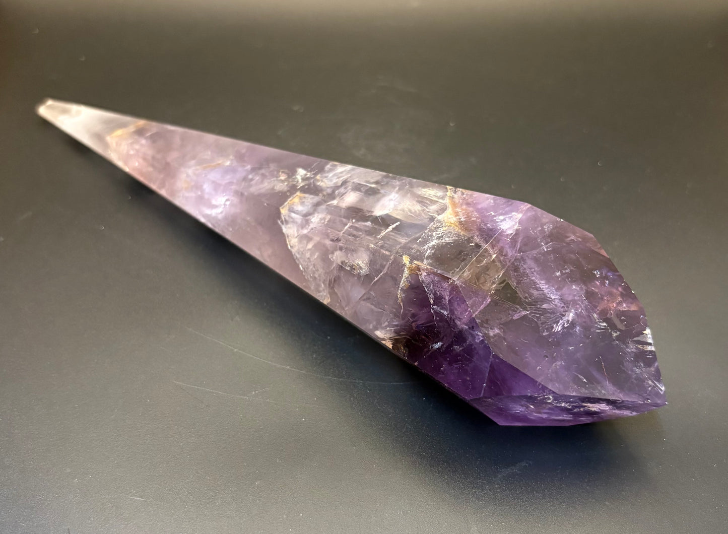 A natural amethyst crystal point tumbler with a pointed tip and faceted bottom, resting on a dark gray surface.