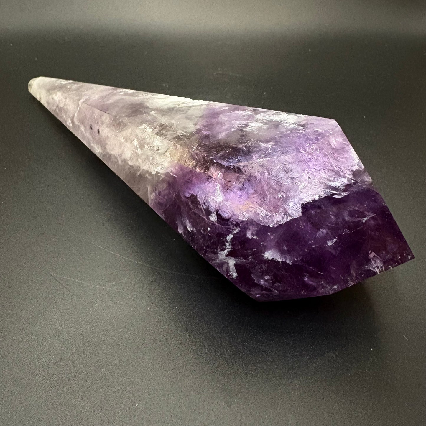 A natural amethyst crystal cone tippic stone with a gradient of purple and white, resting on a dark gray surface.