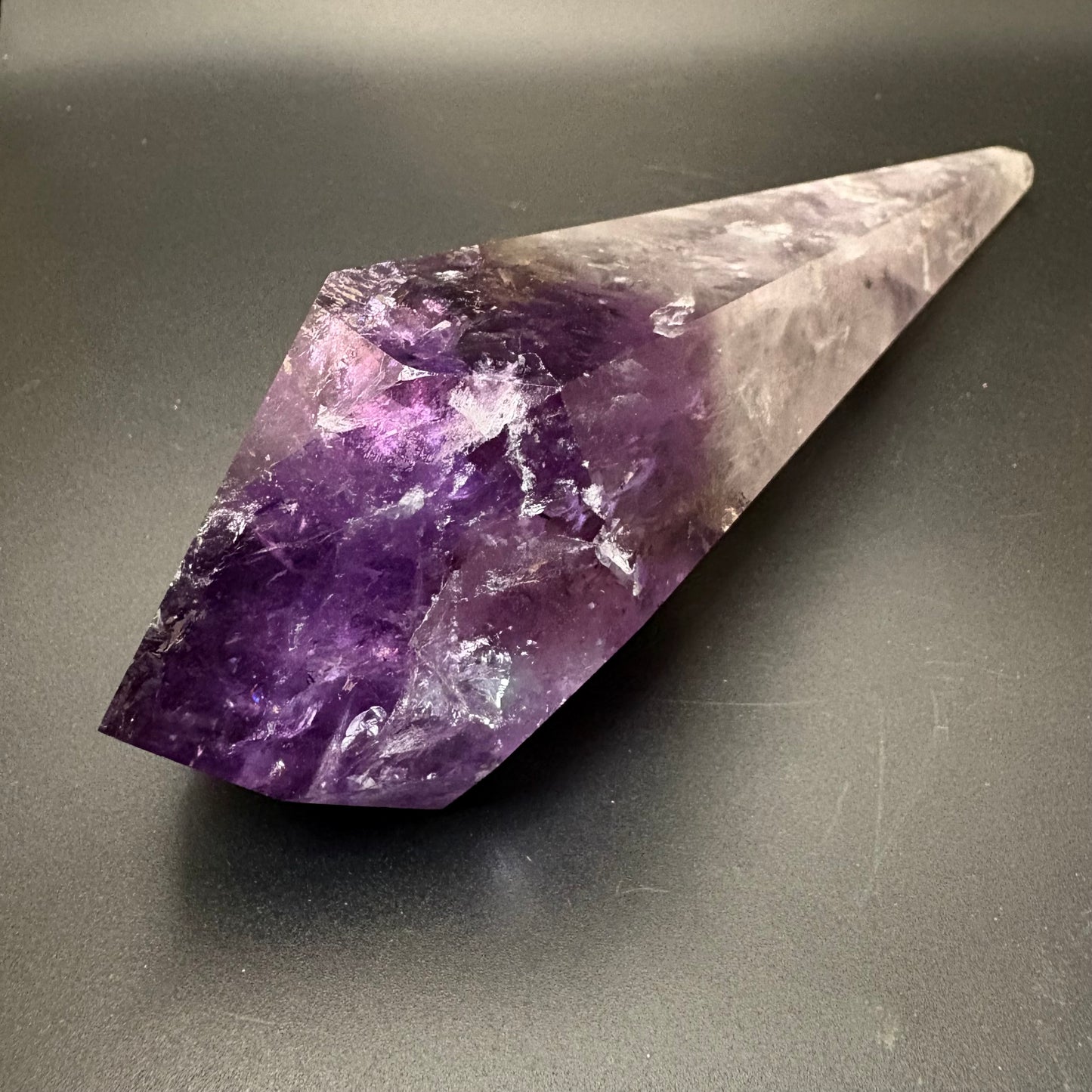 A natural amethyst crystal point with a gradient from deep purple to translucent pale lavender, resting on a dark gray surface.