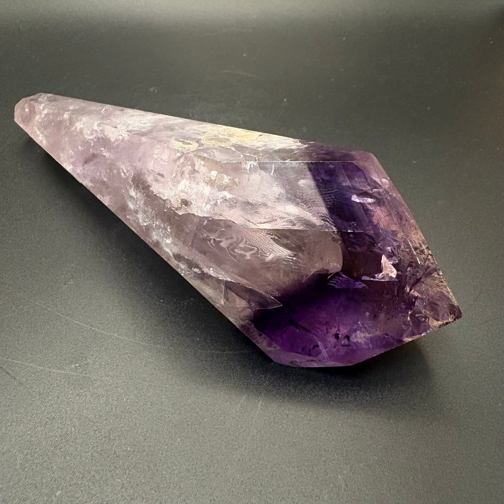 A natural amethyst crystal point tumbler with a gradient from pale lavender to deep purple, resting on a dark gray surface.