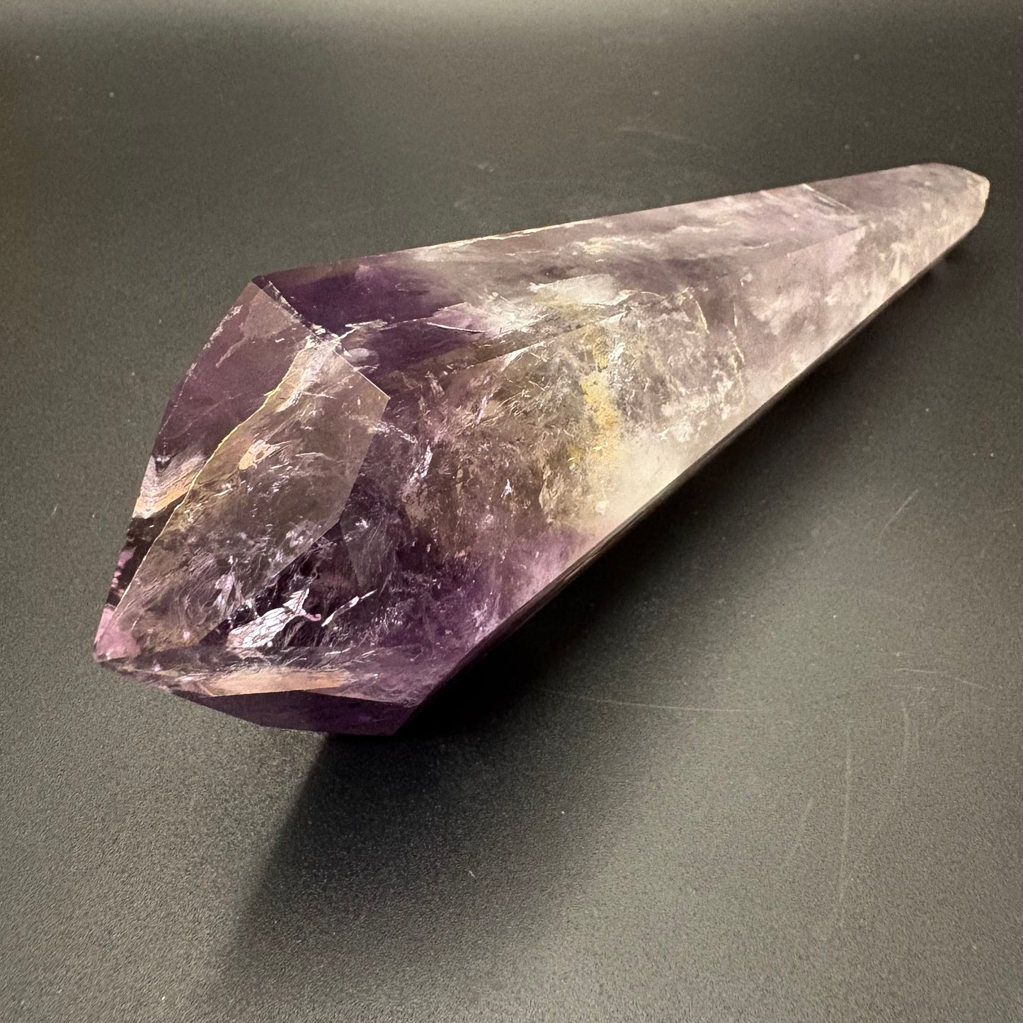 A natural amethyst crystal point with a purple and clear gradient, resting on a dark gray surface.