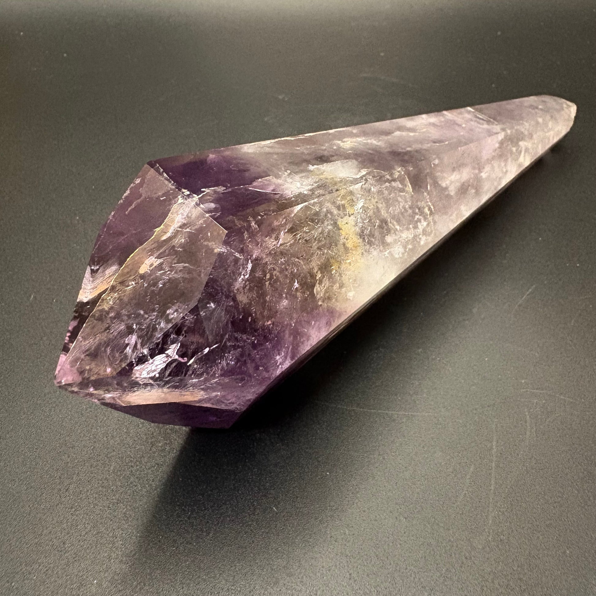 A natural amethyst crystal point with a purple and clear gradient, resting on a dark gray surface.