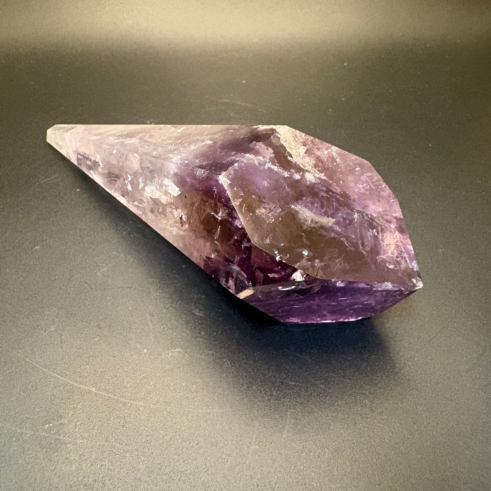 A natural amethyst crystal point with a pointed tip and rough, faceted lower section, resting on a dark gray surface.