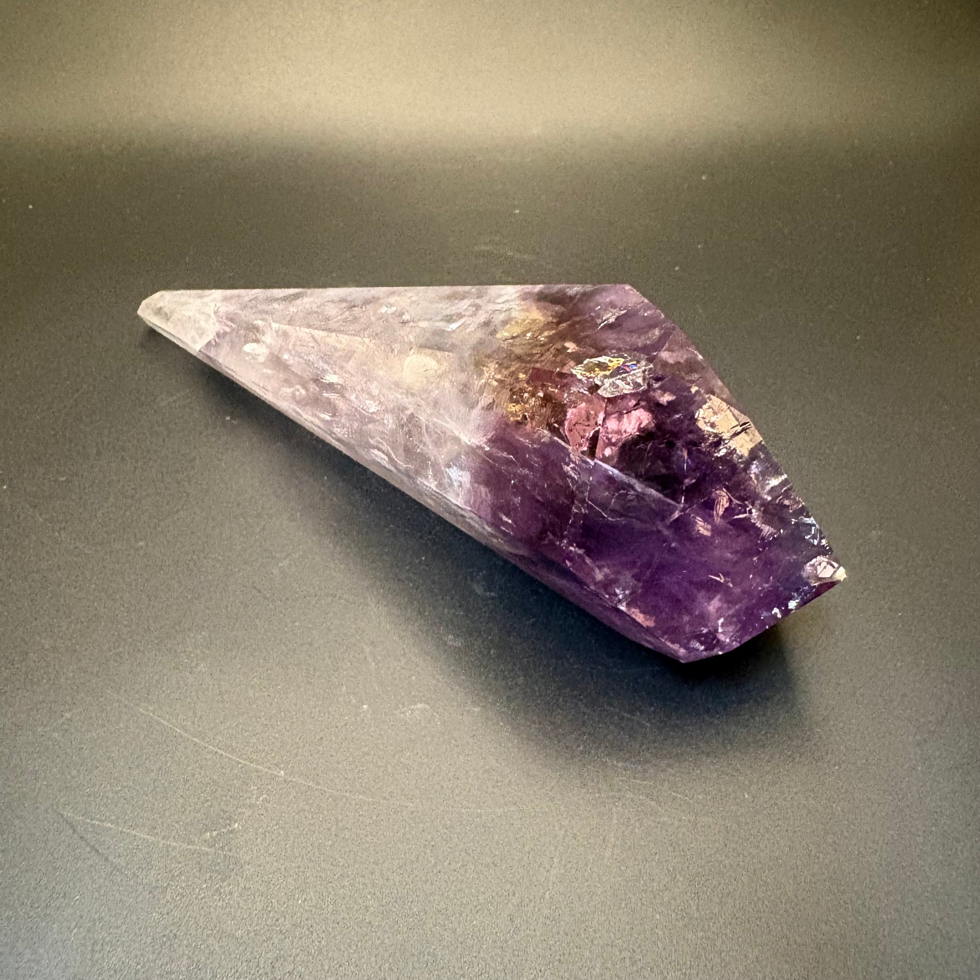 A natural amethyst crystal point with a smooth purple gradient and natural inclusions, resting on a dark gray surface.