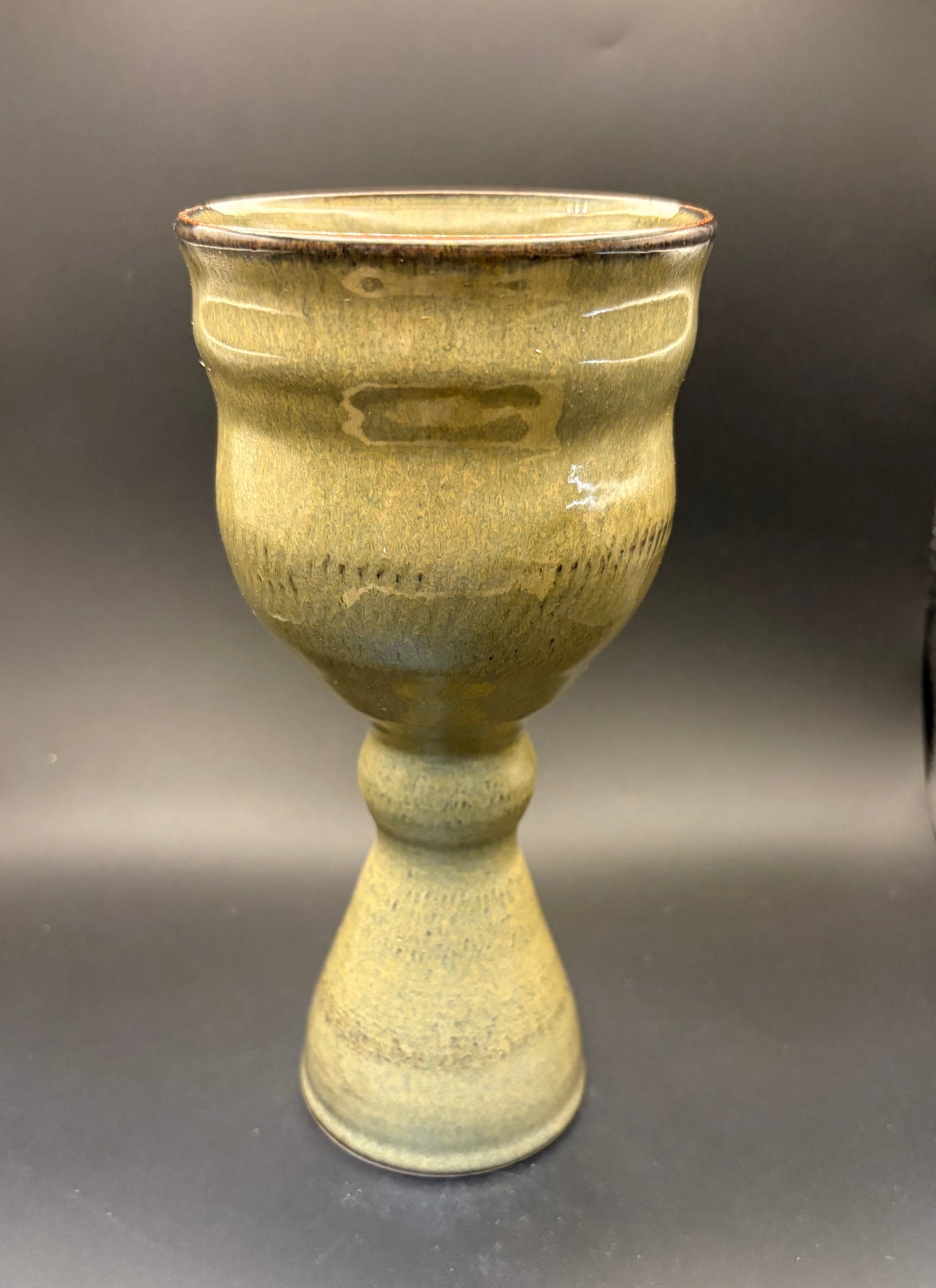 A handmade ceramic goblet or chalice with a textured, ribbed body and a flared base, featuring a rustic olive-green and yellow glaze with a dark brown rim, set against a dark gray background.