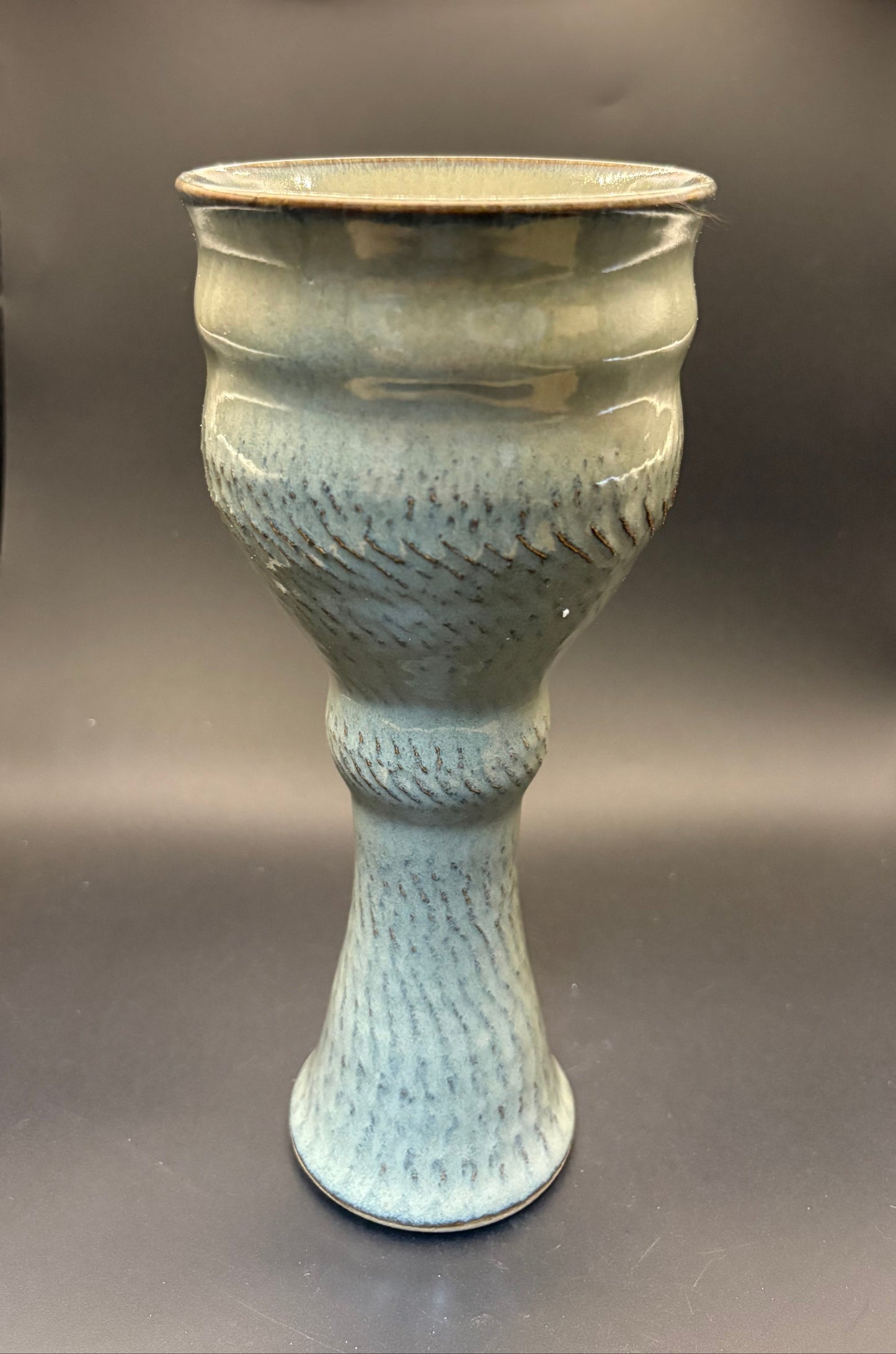 A ceramic goblet or chalice with a textured, ribbed pattern and a glossy pale blue-green glaze, set against a dark gray studio background.