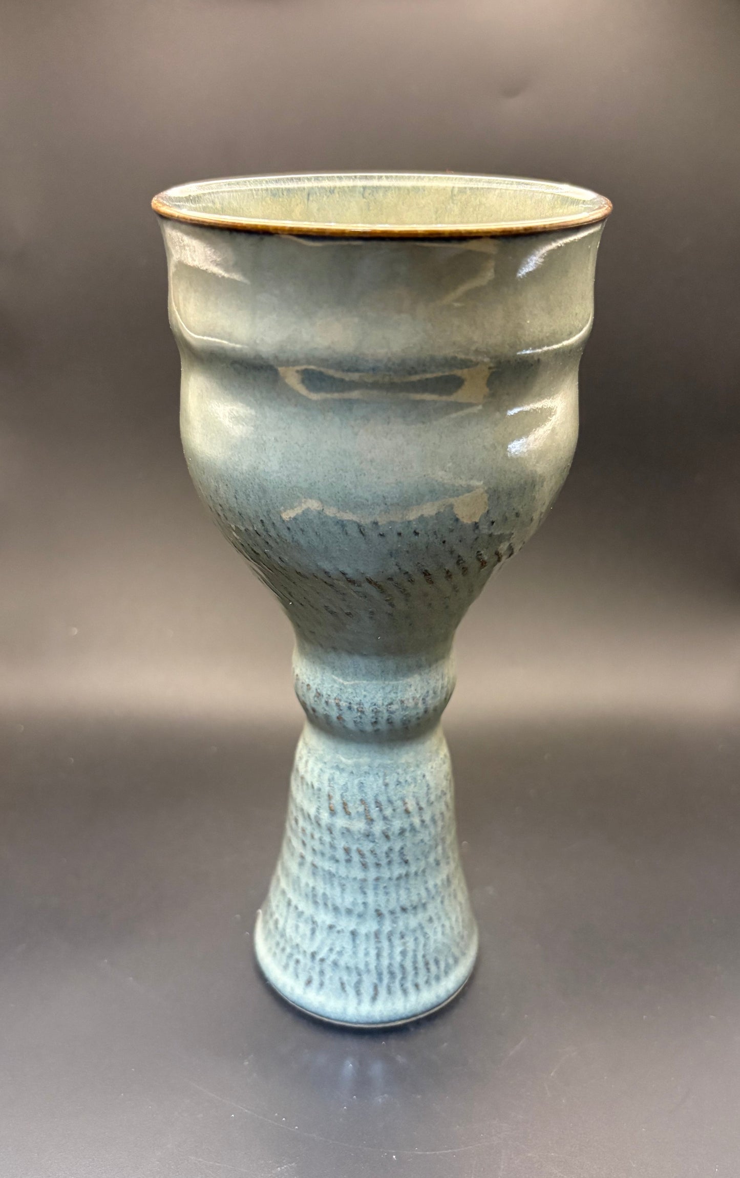 A handmade ceramic goblet with a textured, ribbed foot and a glossy light blue-green glaze, set against a dark gray studio background.