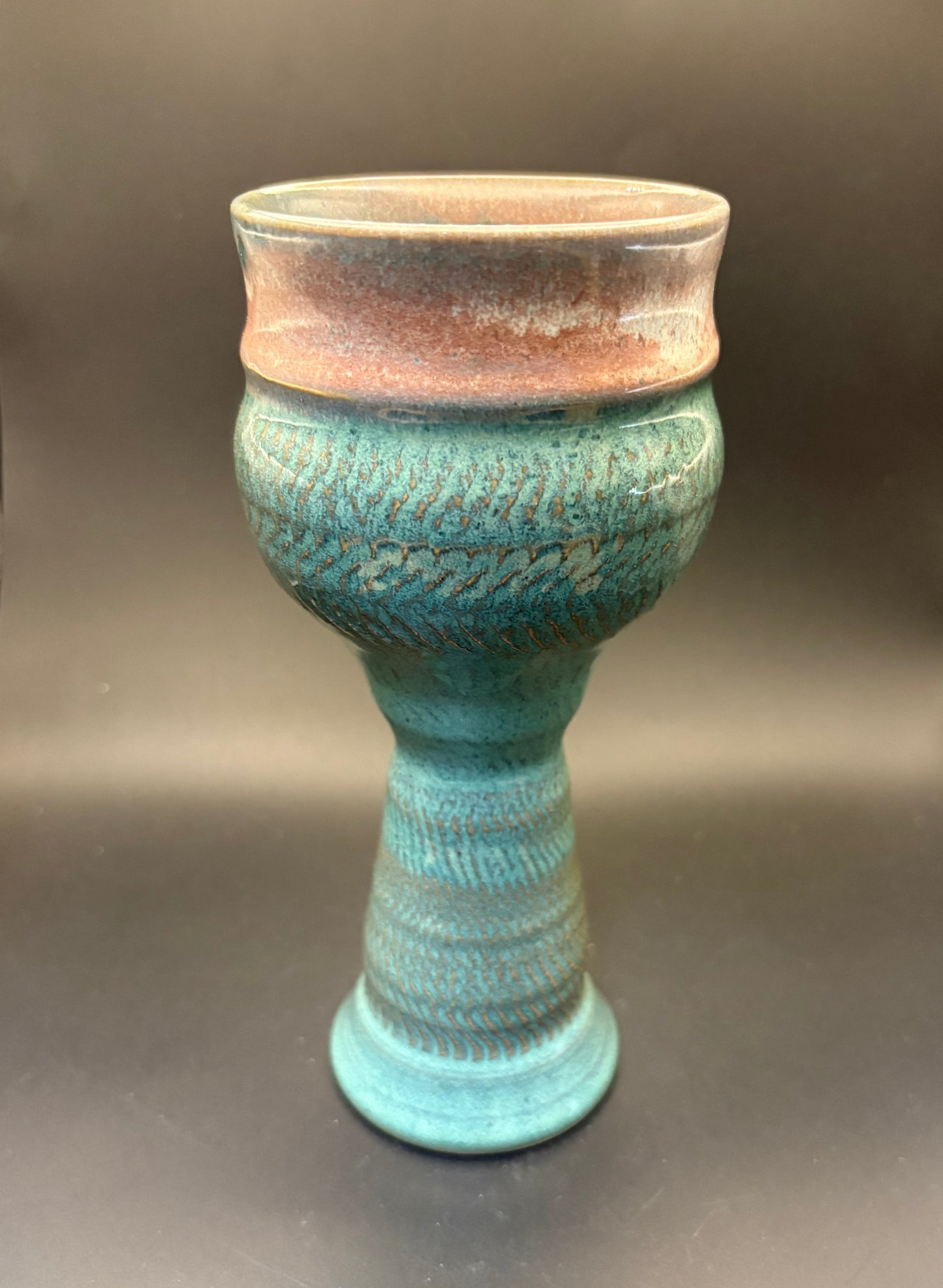 A handmade ceramic goblet or cup with a textured turquoise glaze and a contrasting terracotta-colored rim, set against a dark gray background.