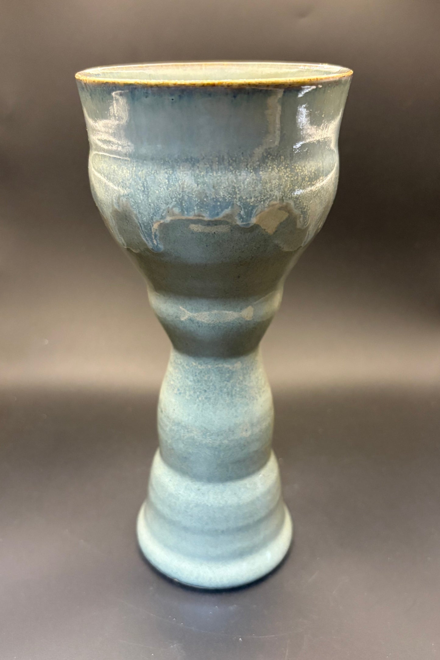 A handmade ceramic goblet with a textured, ribbed design and a glossy glaze in shades of blue-green and gray, featuring a rustic gold-brown rim, set against a dark gray background.