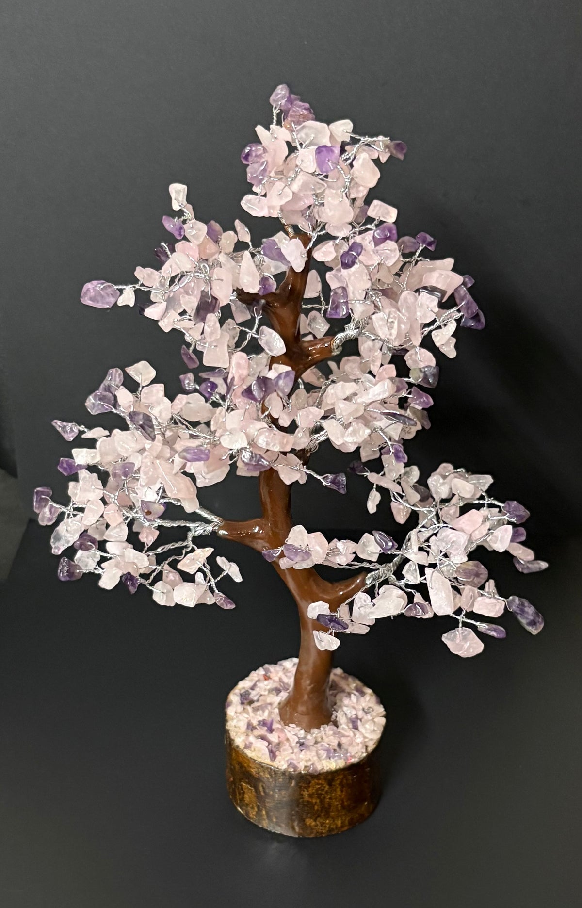 Crystal tree sculpture with rose quartz and purple amethyst leaves on a brown branch structure, set in a circular stone base against a dark gray background.