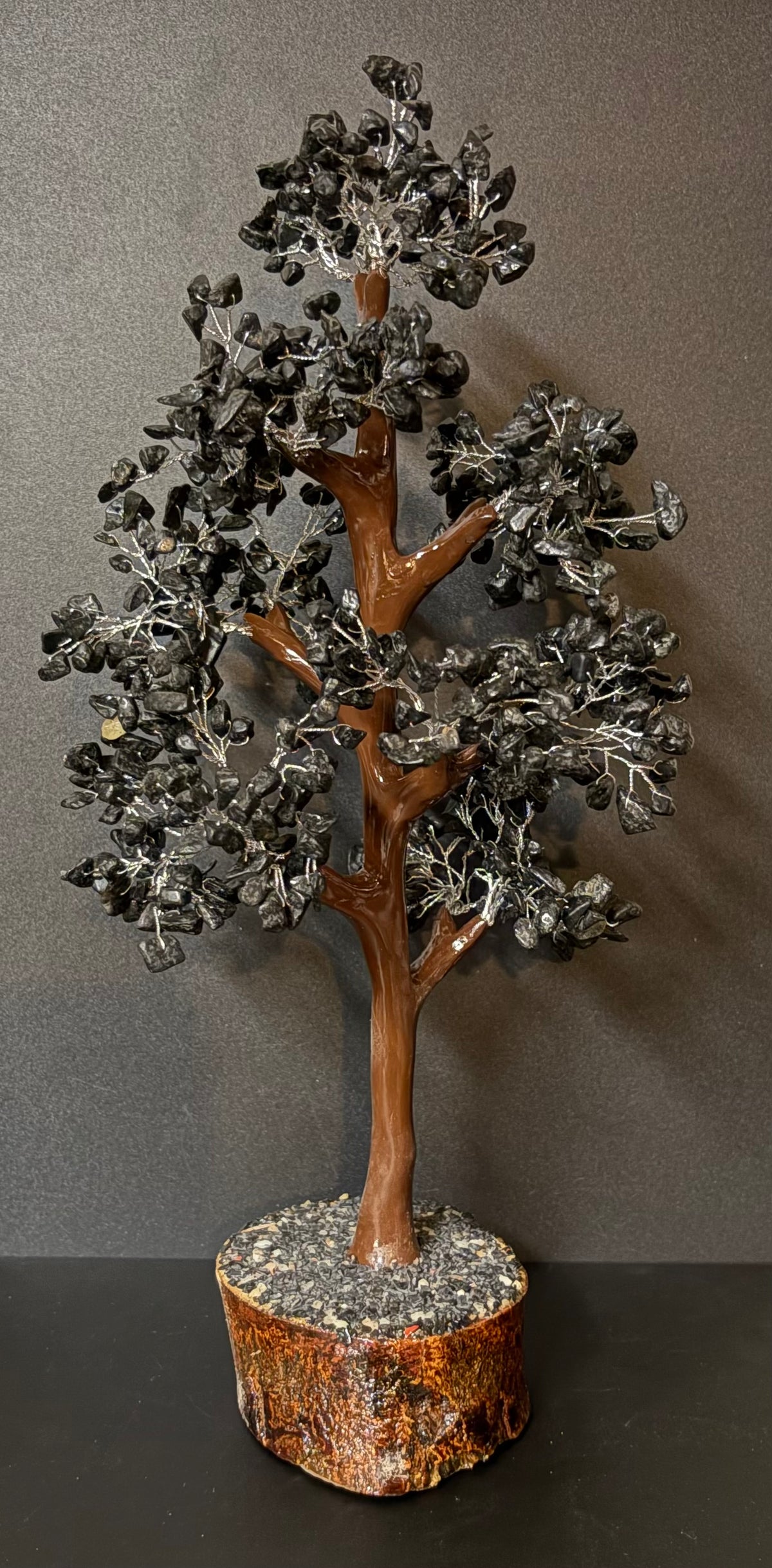 A crystal tree sculpture made of polished black tourmaline leaves and branches with a brown tree trunk, mounted on a round polished stone base, set against a dark gray background.