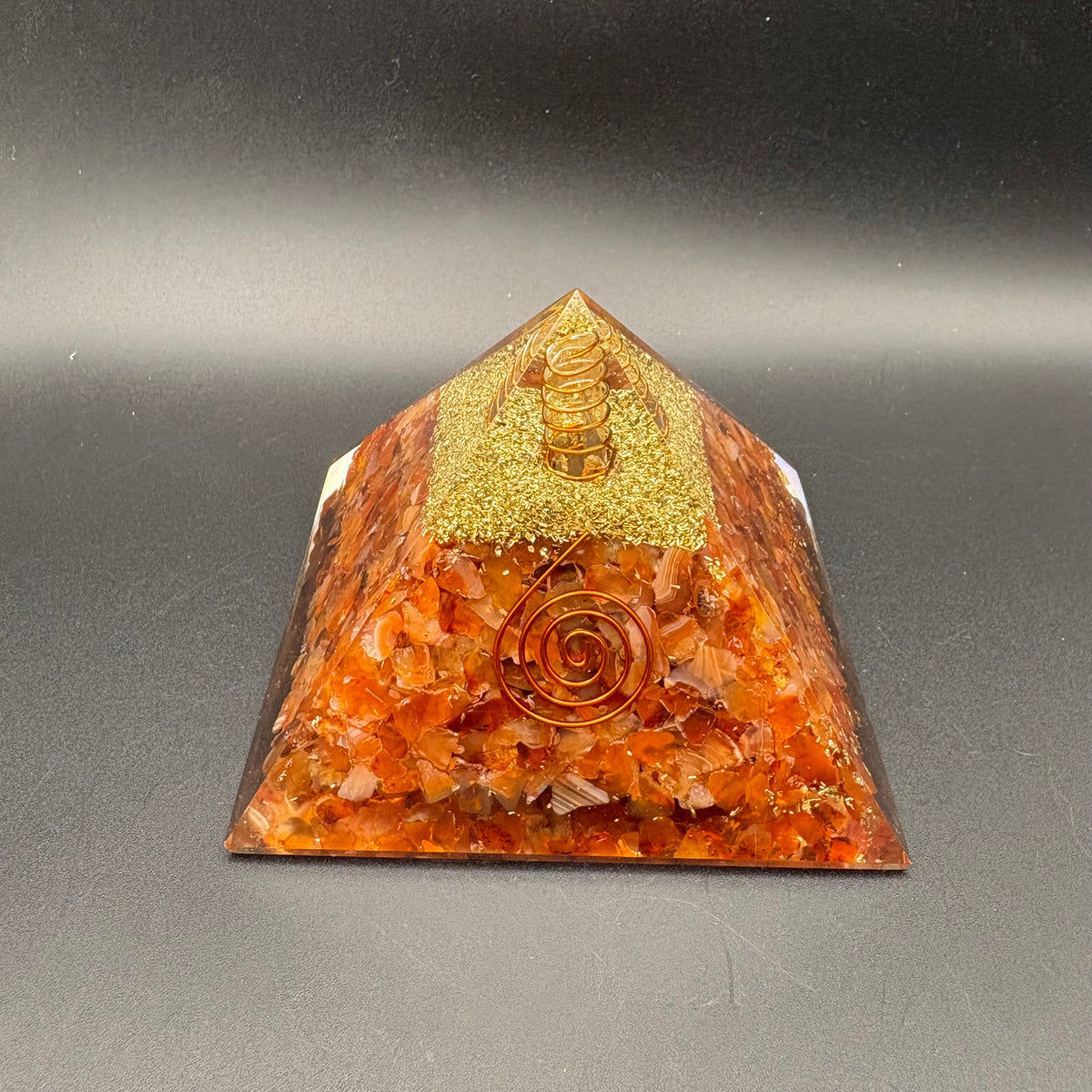 An orange and gold organic resin pyramid chakra statue filled with crushed Carnelian Crystals, featuring a gold glitter top section and a central spiral design, set against a dark gray background.